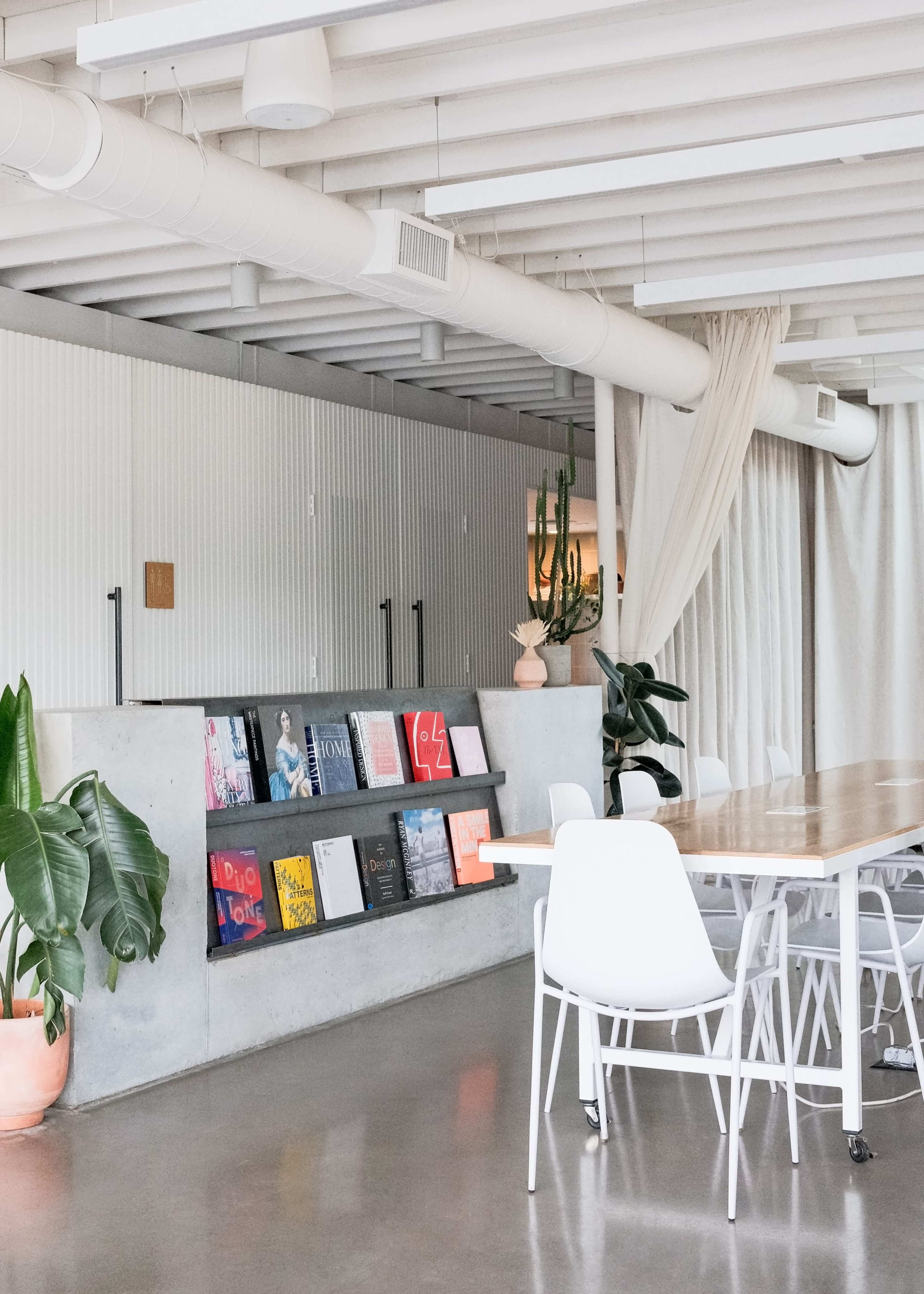 A modern workspace features a long wooden table surrounded by white chairs, with a display of colorful books arranged on a concrete shelf and potted plants nearby.