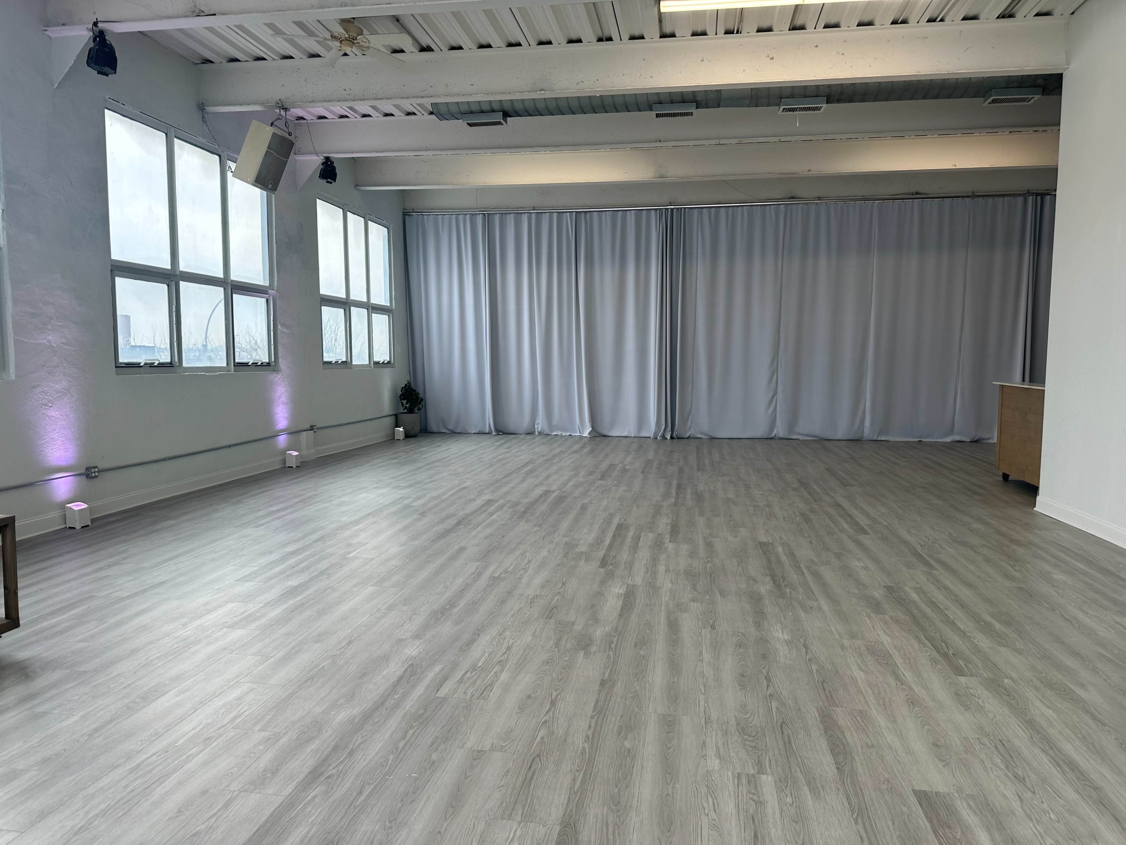 The image shows an empty room with large windows, light-colored flooring, and gray curtains at one end.