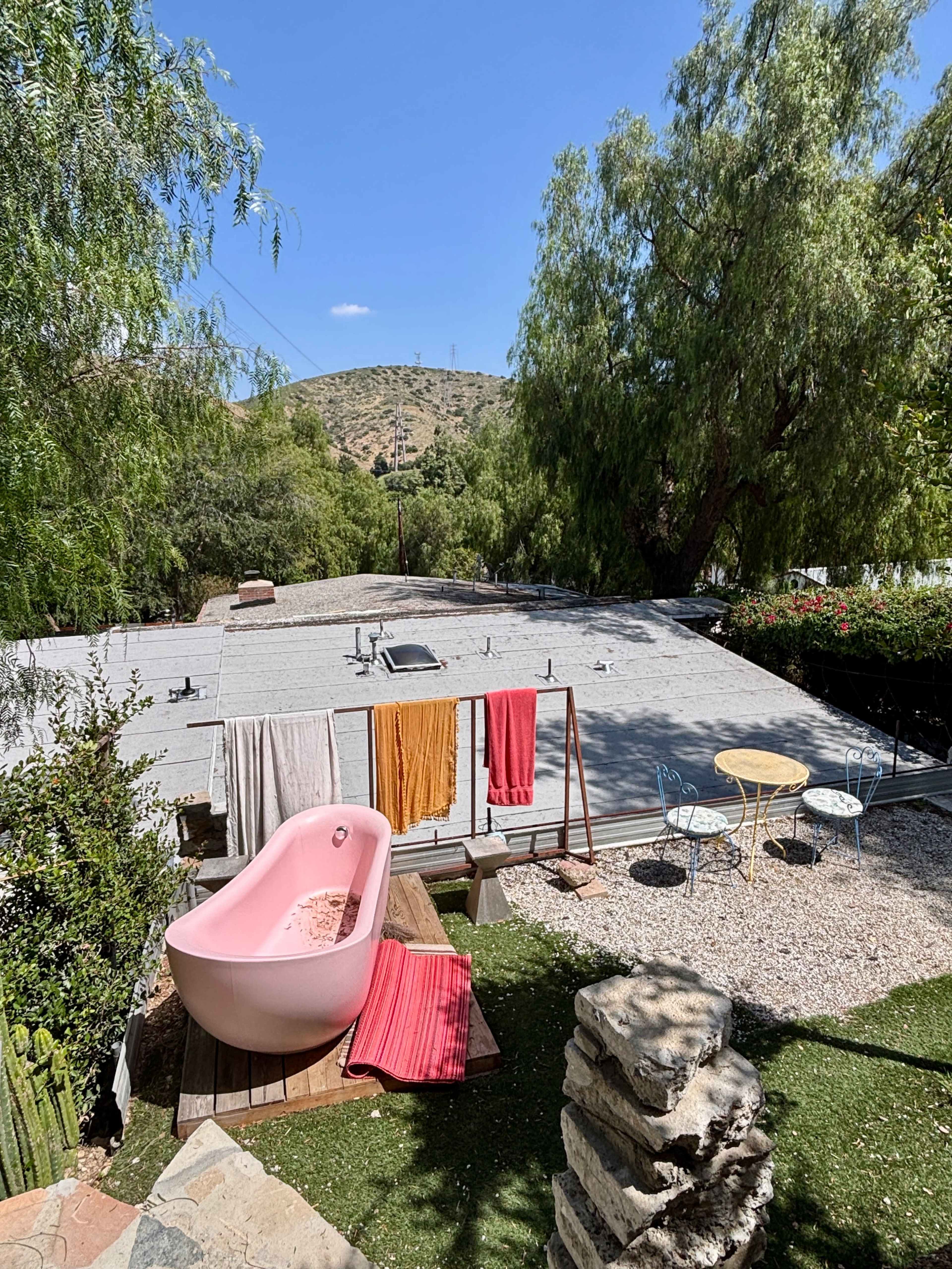 The image shows a pink bathtub on a grassy area surrounded by colorful towels hanging on a clothesline, with a small table and chairs in the foreground and a hillside in the background.