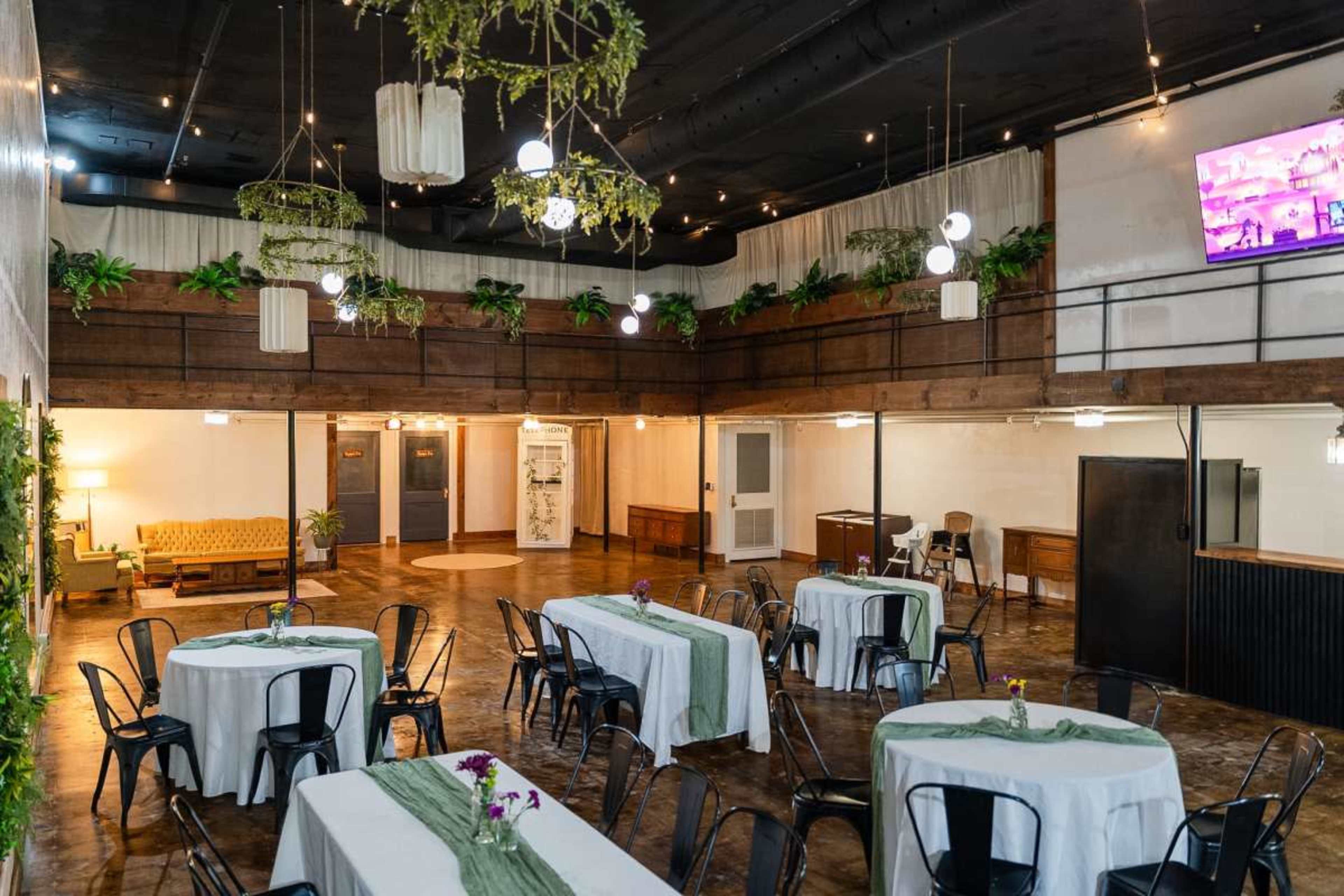 The image shows a spacious indoor event venue featuring several tables with white tablecloths, greenery decor, and a mix of wooden and metal furnishings.