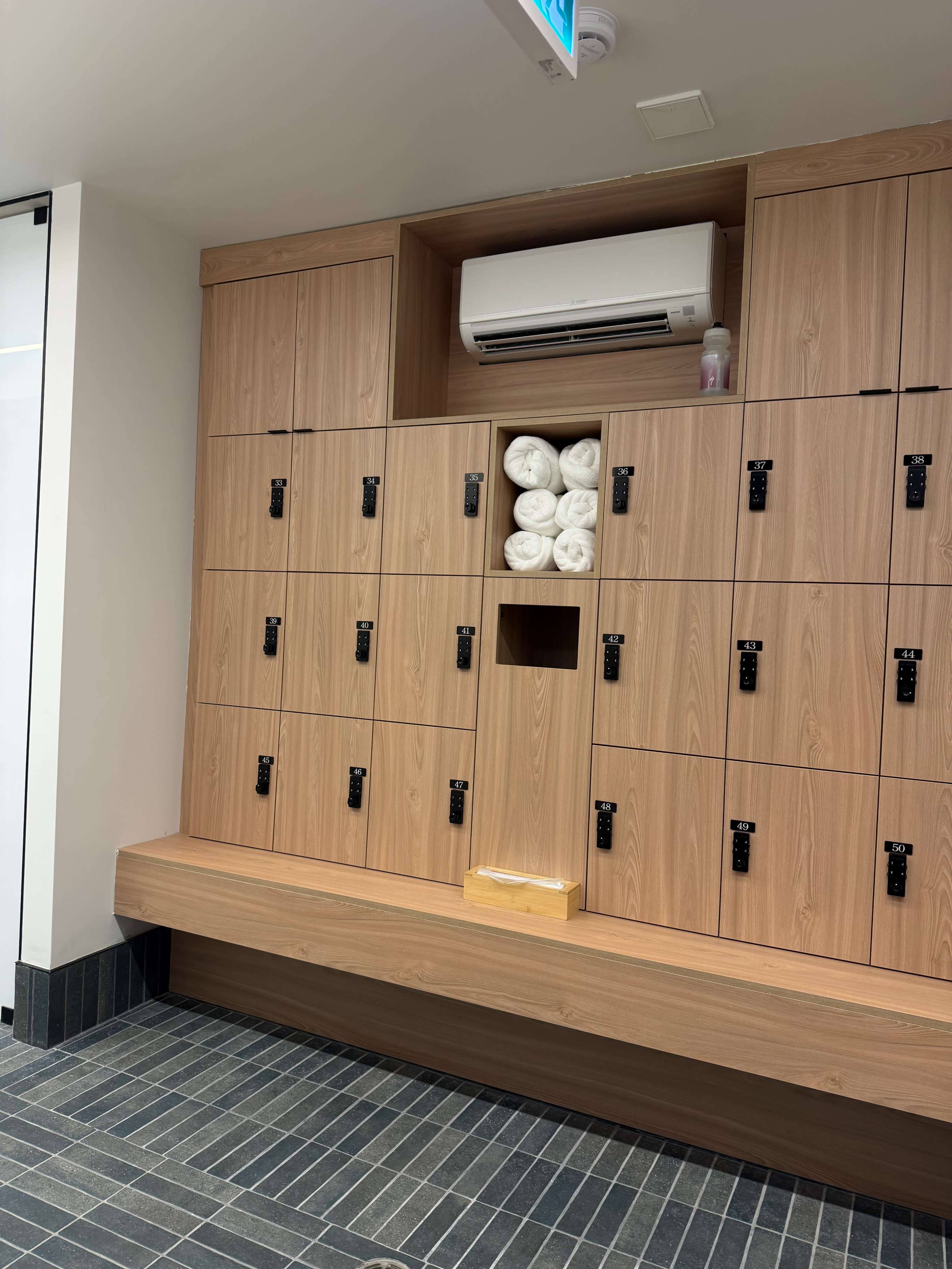 The image shows a row of wooden lockers, featuring some stacked towels and an air conditioning unit above them.