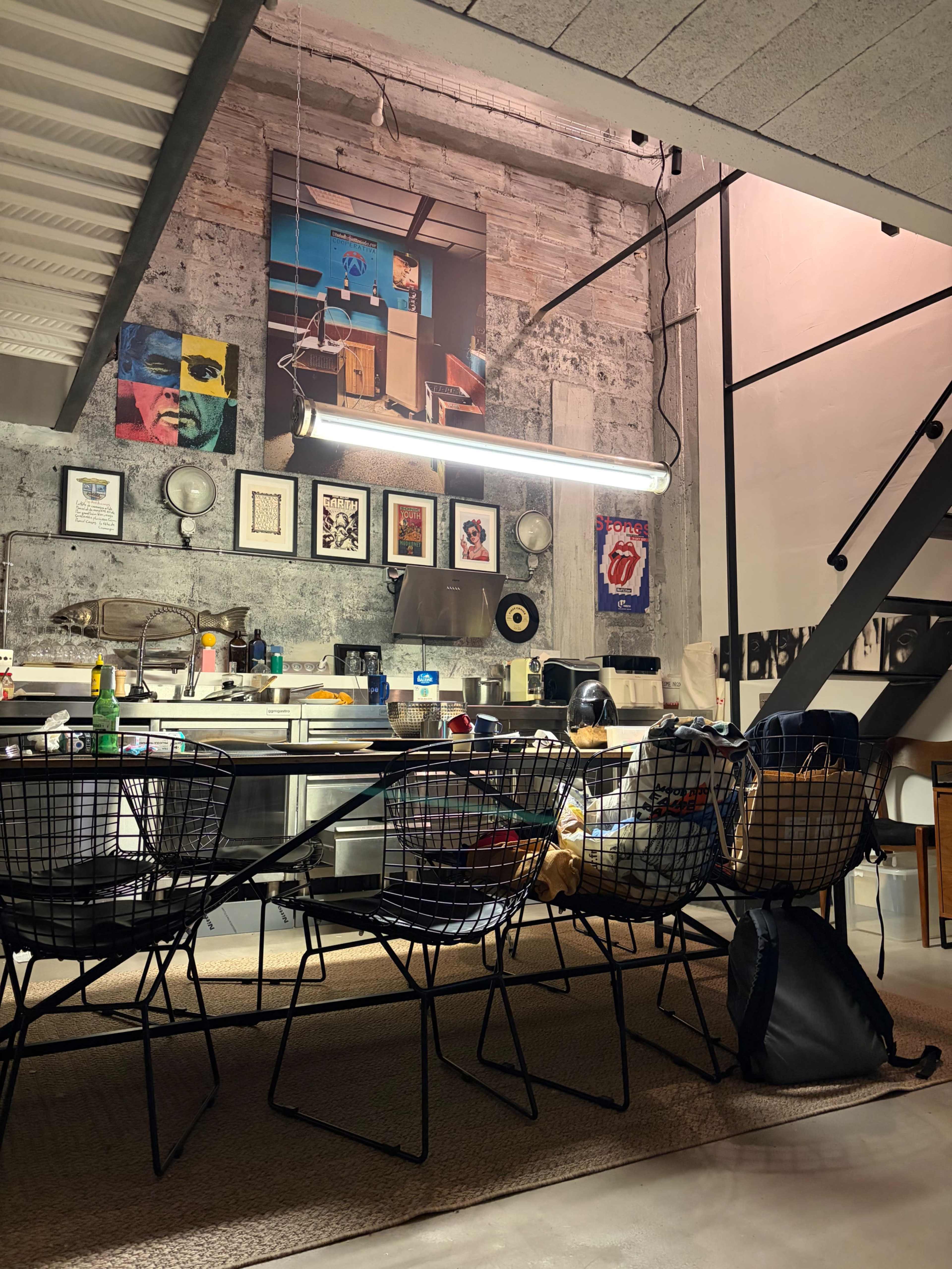 The image shows a modern kitchen and dining area with metal chairs, a concrete wall adorned with artwork, and a staircase leading to an upper level.