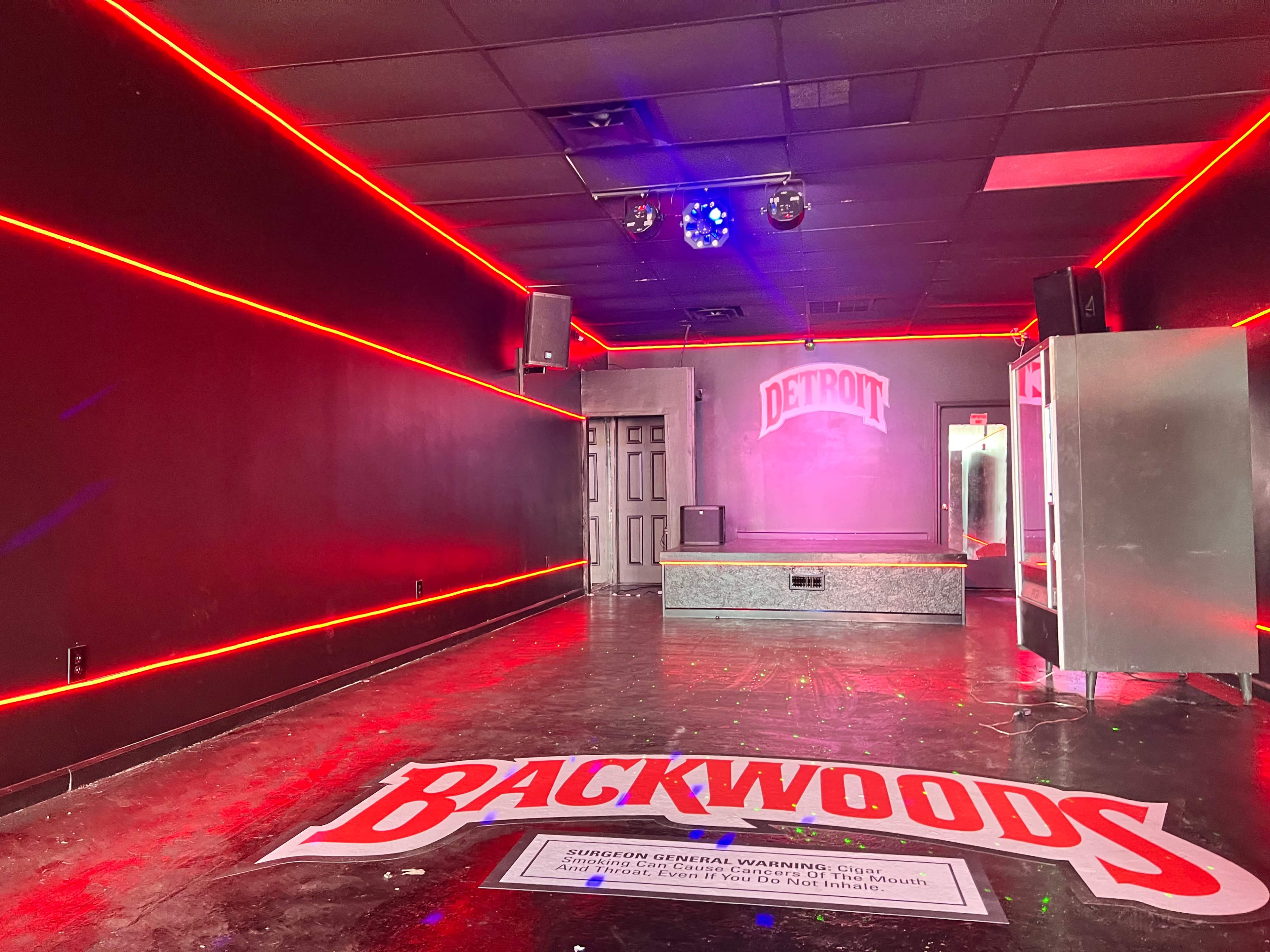 An interior space with red neon lighting, a small stage, and a large banner on the floor displaying the word "BACKWOODS."