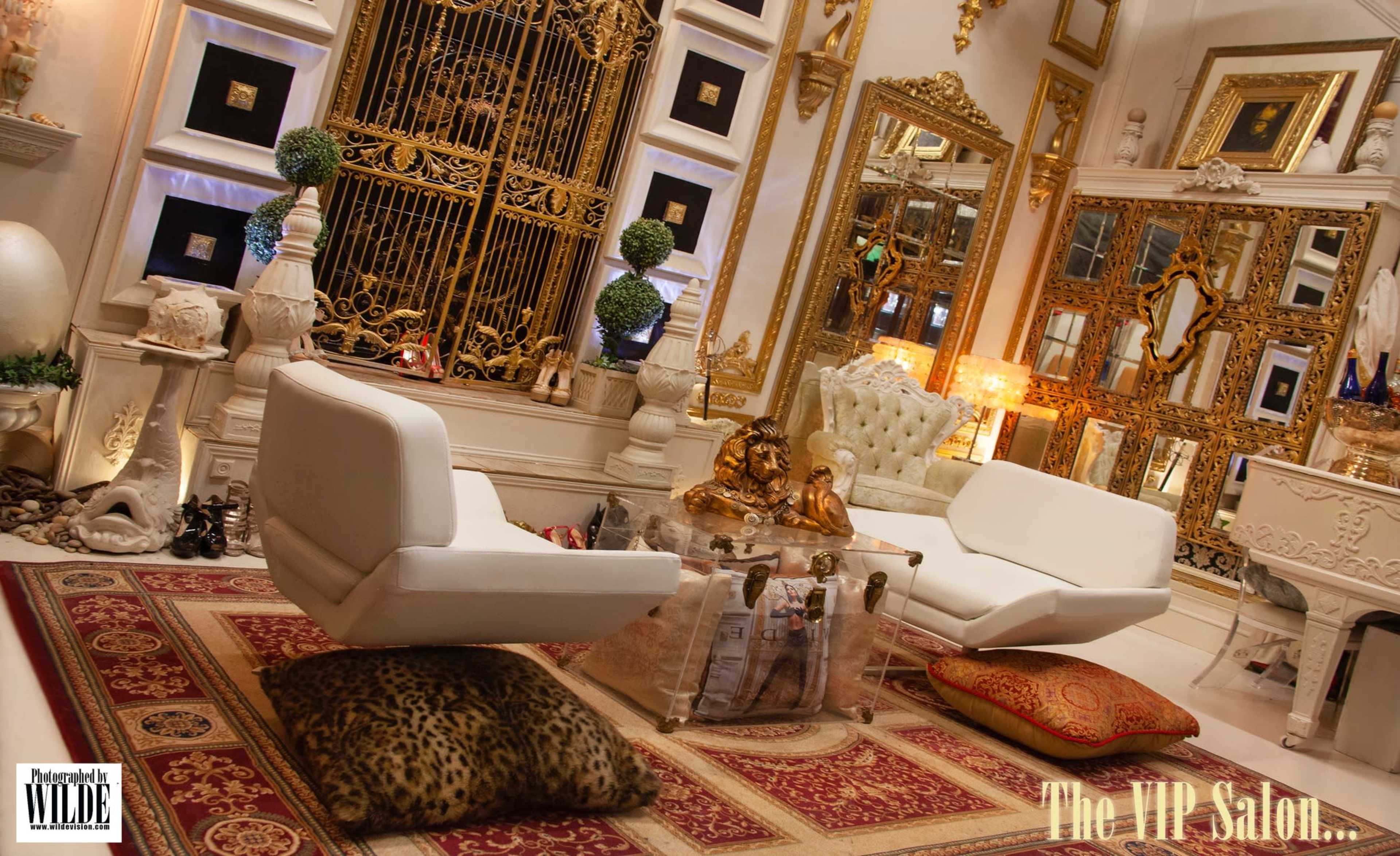 The image depicts an opulent interior design of a salon featuring two white chairs, a glass coffee table, and ornate gold mirrors on the walls.