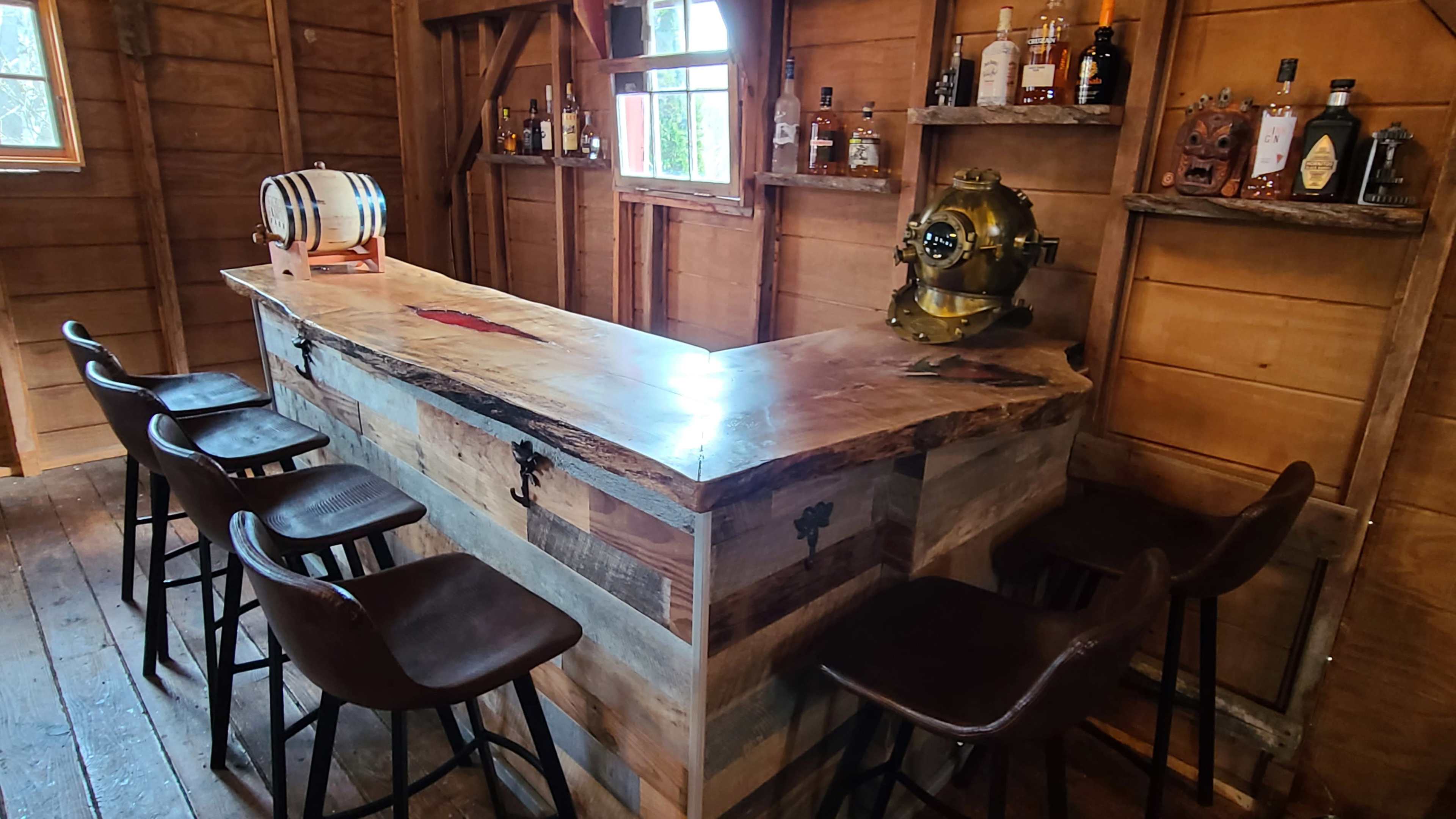 A wooden bar with a rustic finish, surrounded by high-backed stools, inside a cozy cabin-like interior featuring shelves with various bottles.