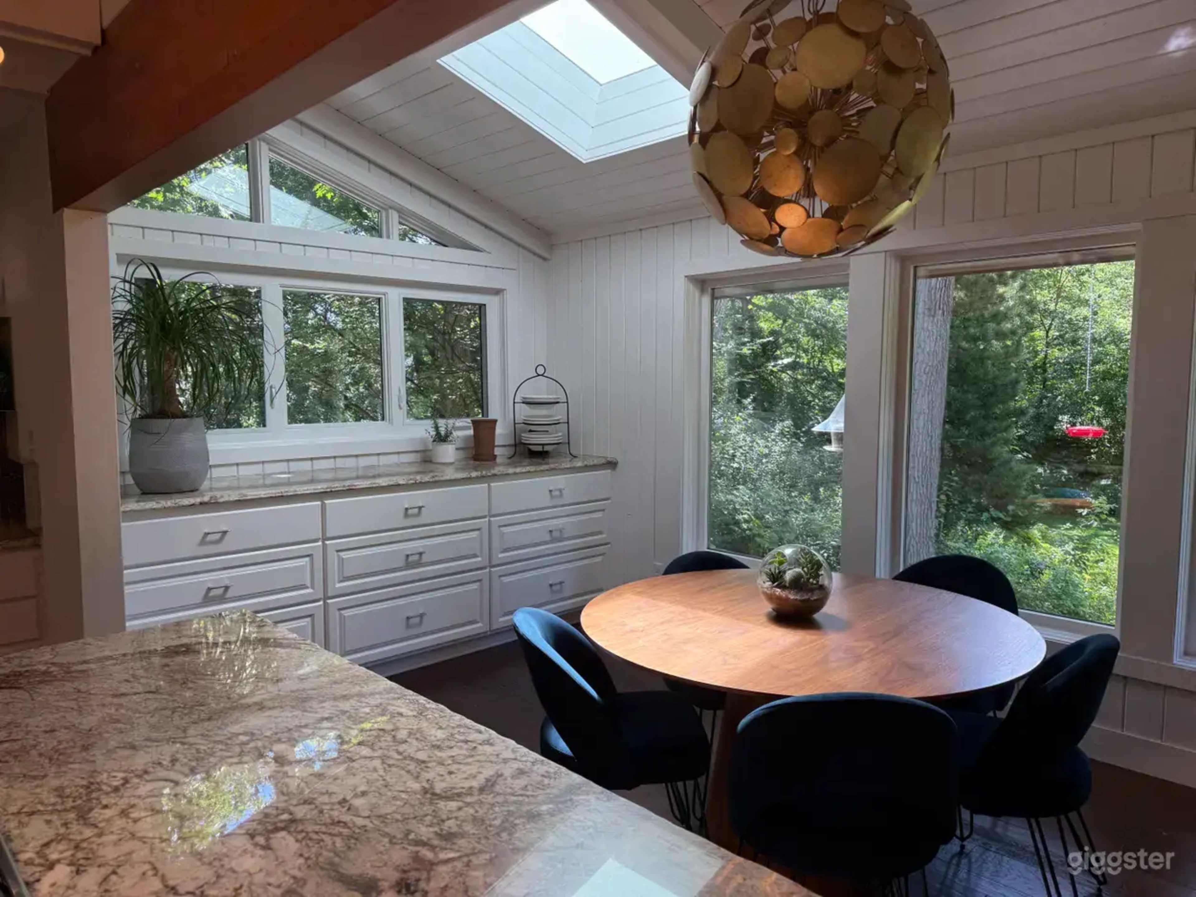 A modern kitchen features a round wooden table surrounded by black chairs, large windows letting in natural light, and a pendant light hanging from the ceiling.