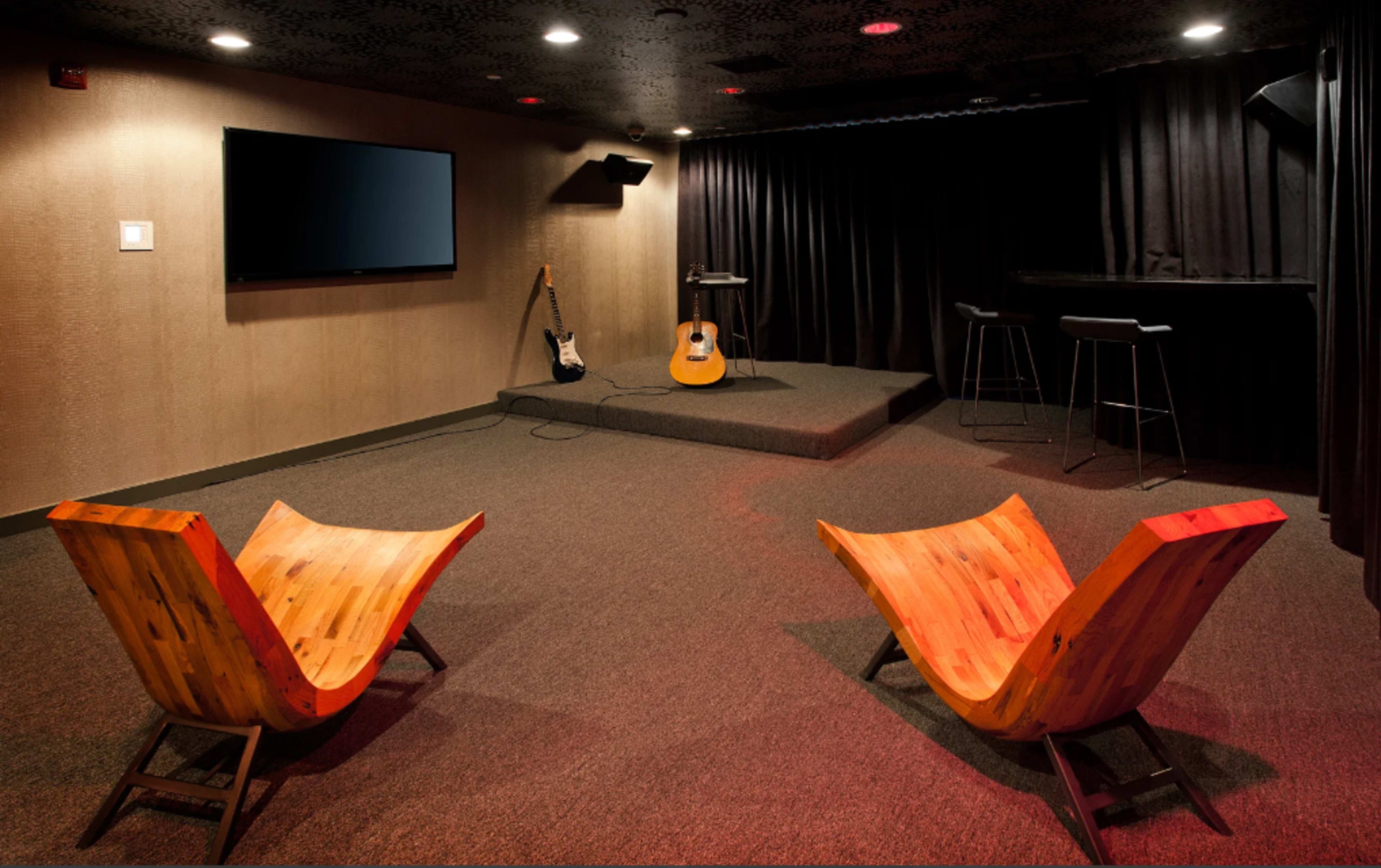The image shows a small performance space with two wooden chairs facing a stage equipped with a guitar, microphone stand, and a TV screen.