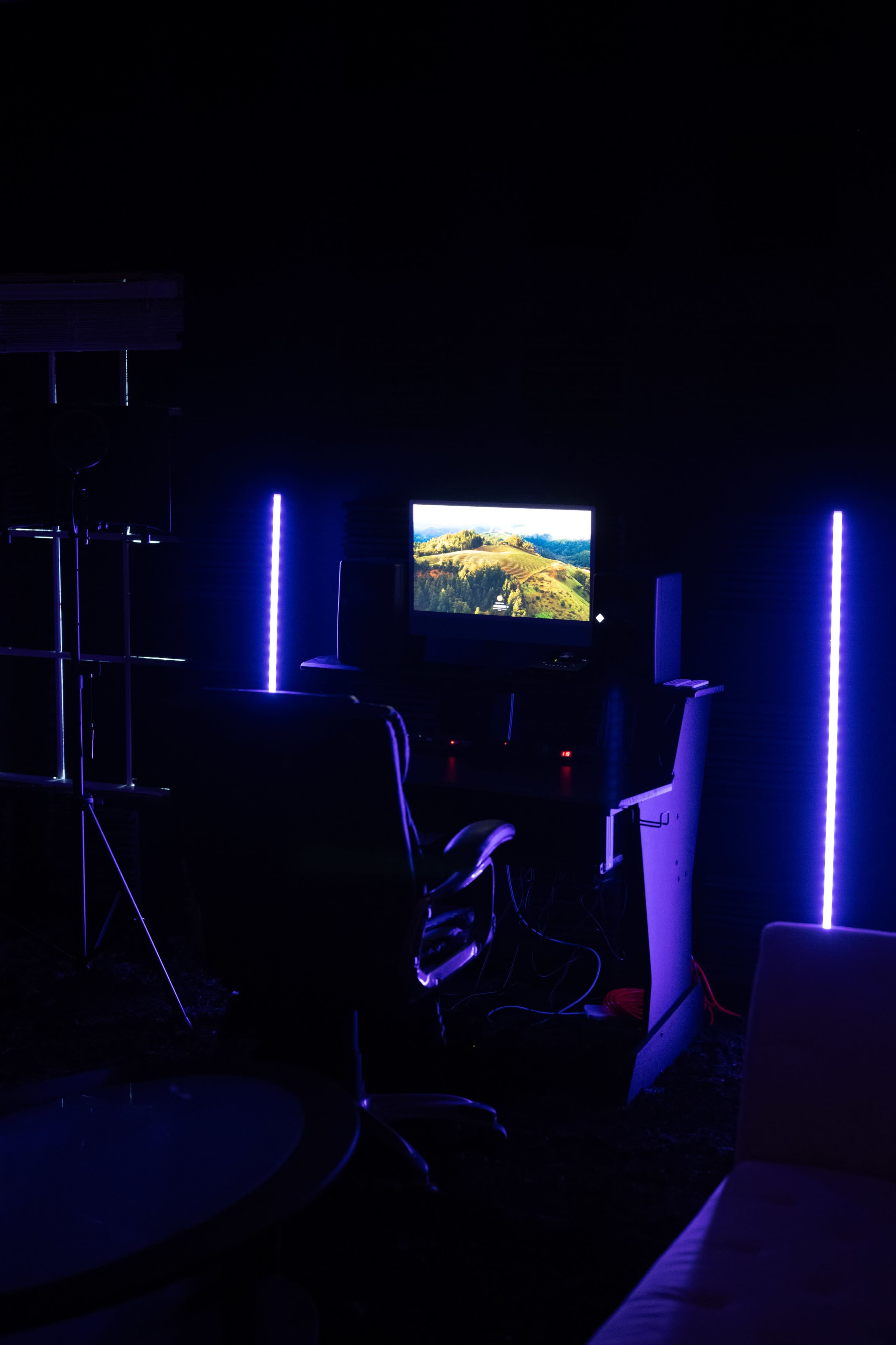 A dark room featuring a computer desk with a monitor displaying a landscape, illuminated by purple LED lights.