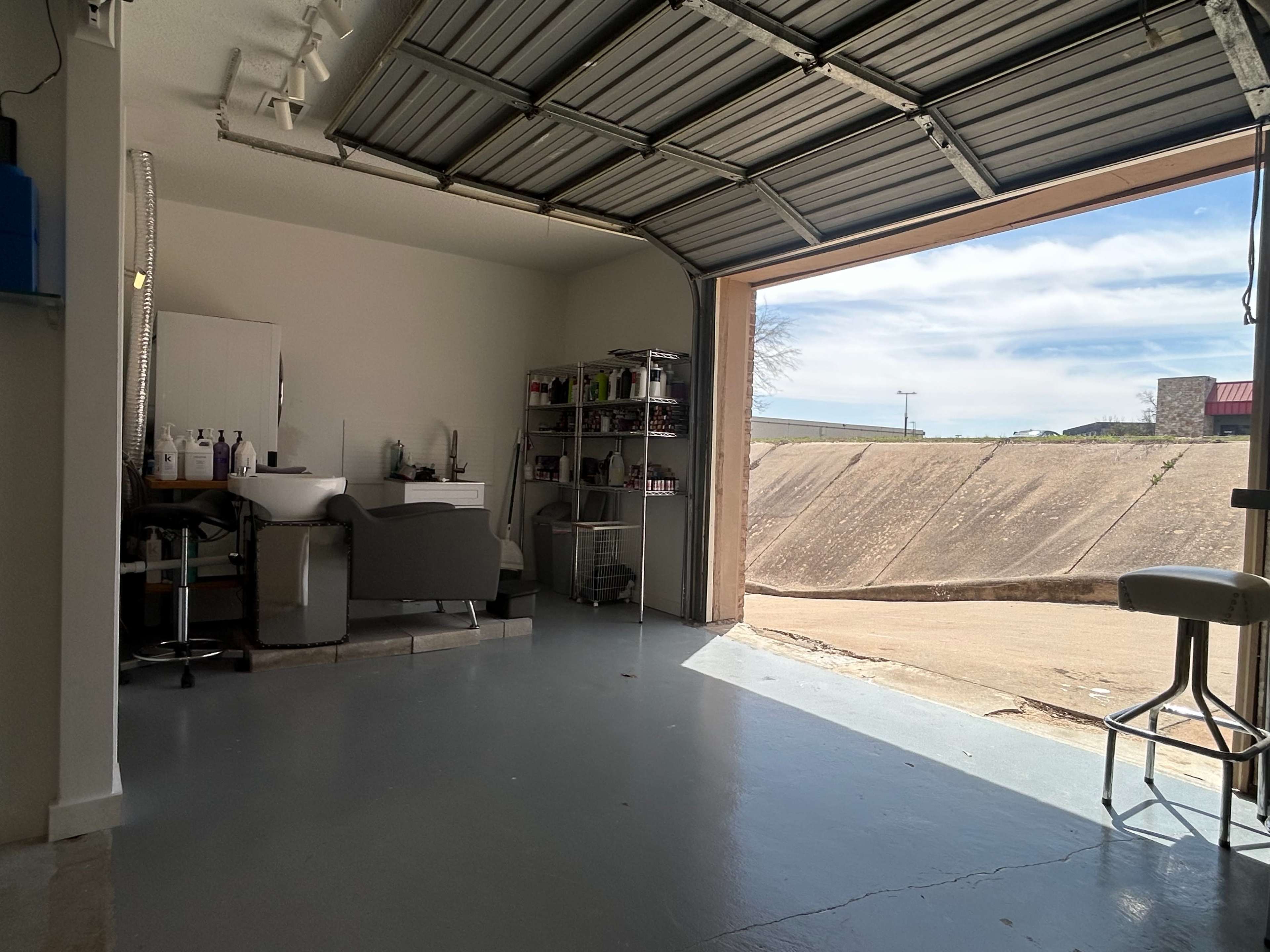 A garage with an open door revealing a concrete driveway and shelves containing various items inside.