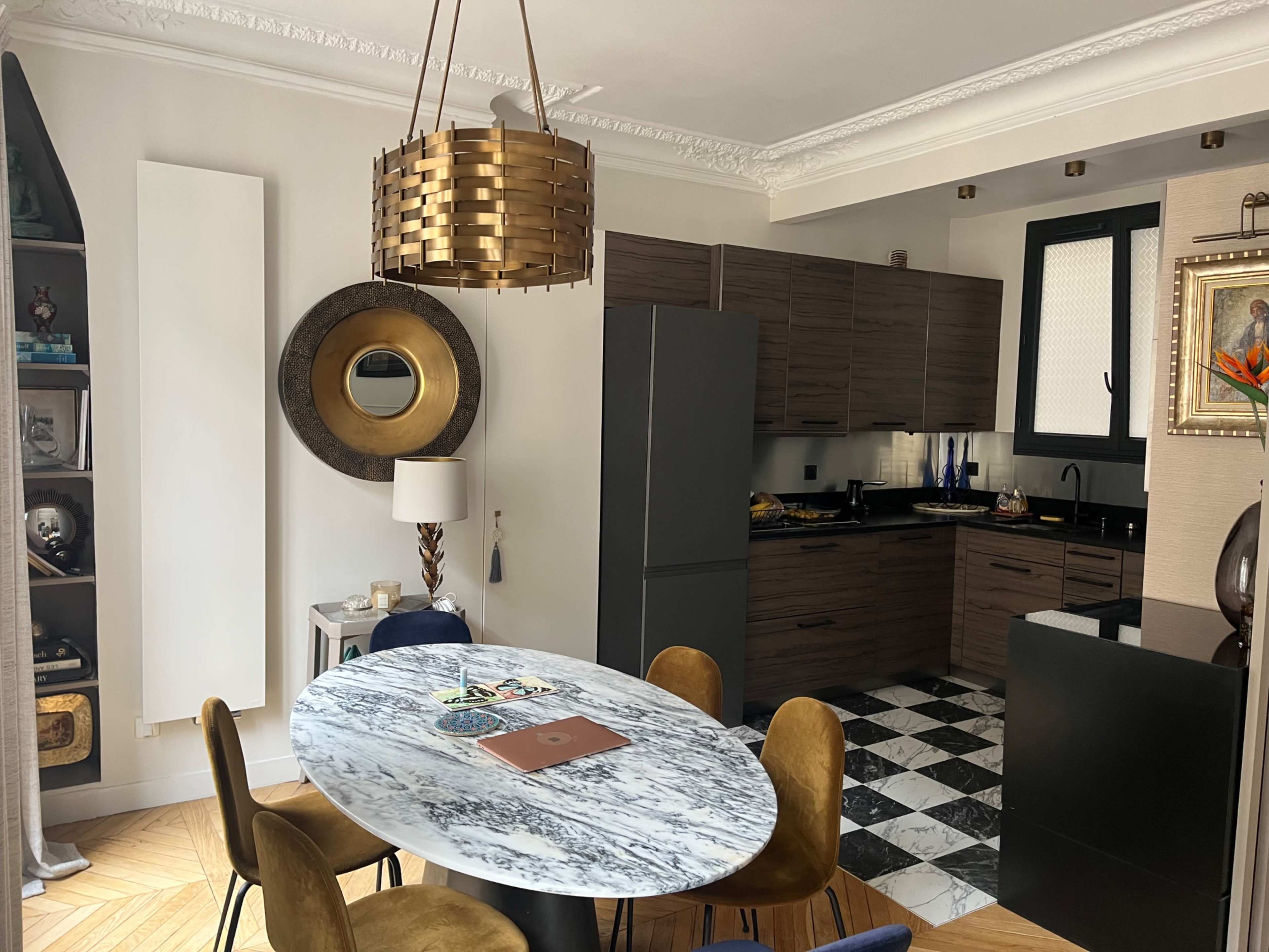 The image shows a modern kitchen and dining area featuring a round marble table, wooden chairs, and sleek cabinetry, with a checkered floor and decorative elements on the walls.