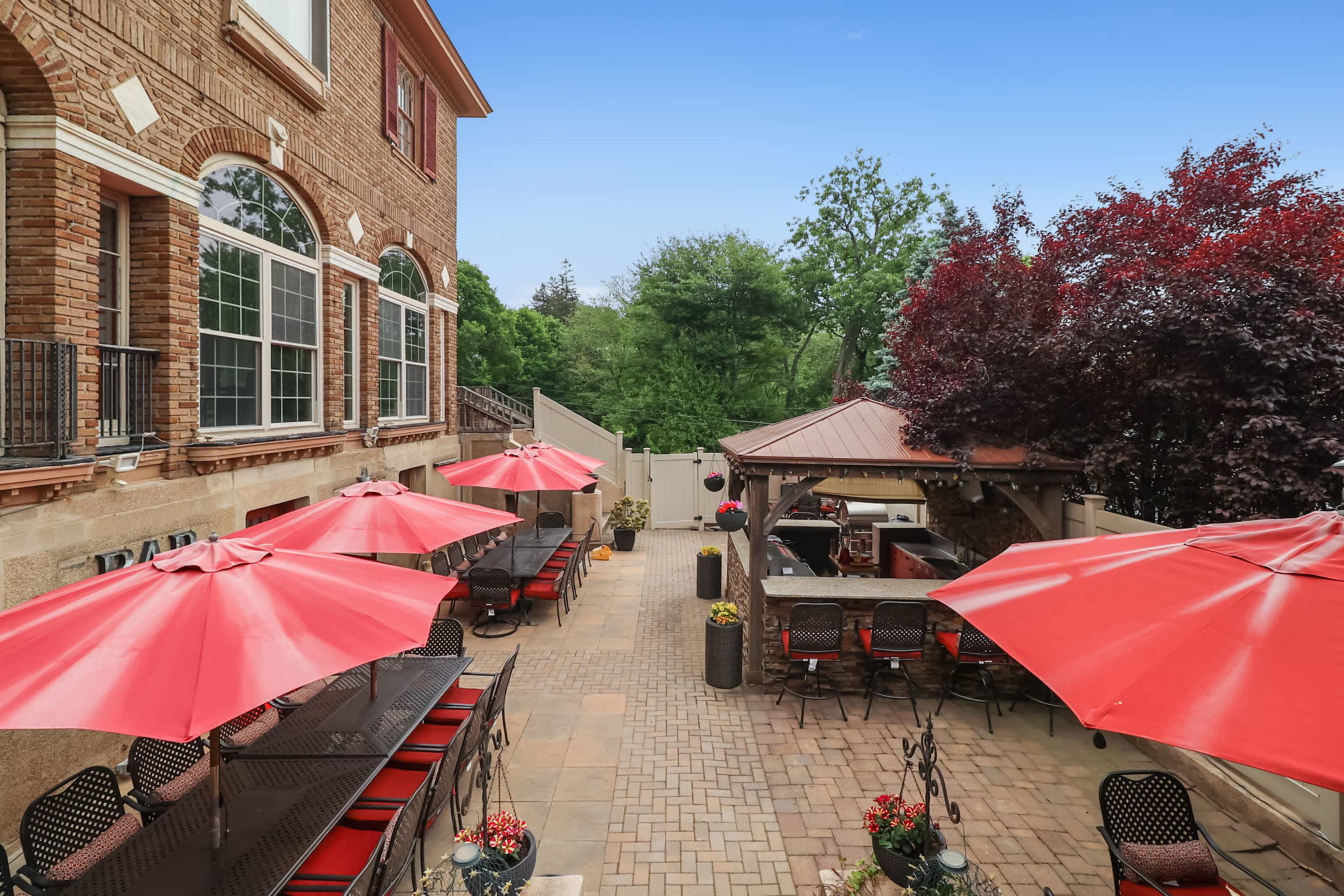 A brick-paved patio features red umbrellas over metal tables and chairs, with a gazebo and trees in the background.