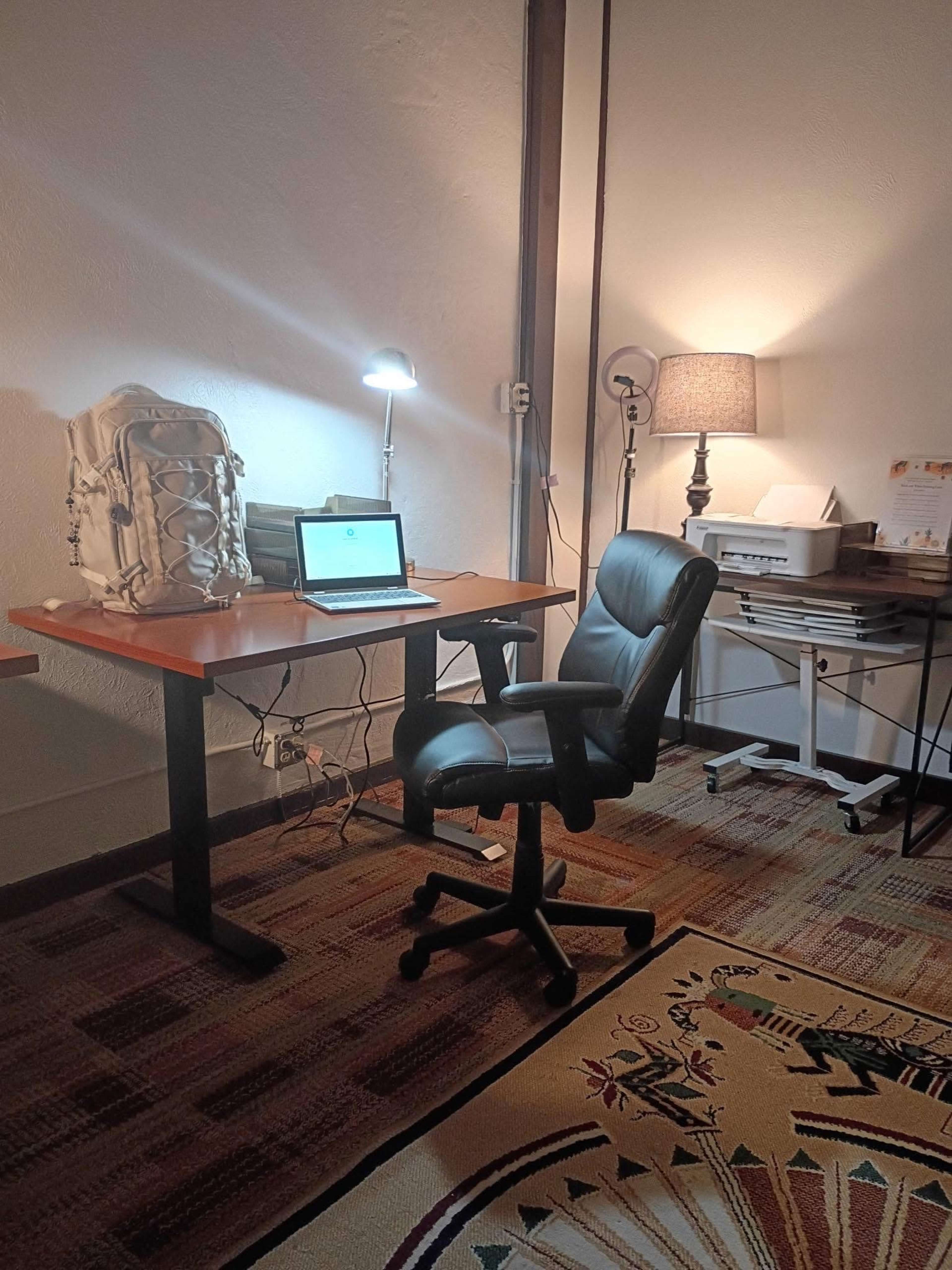 A workspace features a desk with a laptop, a desk lamp, a black swivel chair, and a backpack, along with a side table that has a lamp.