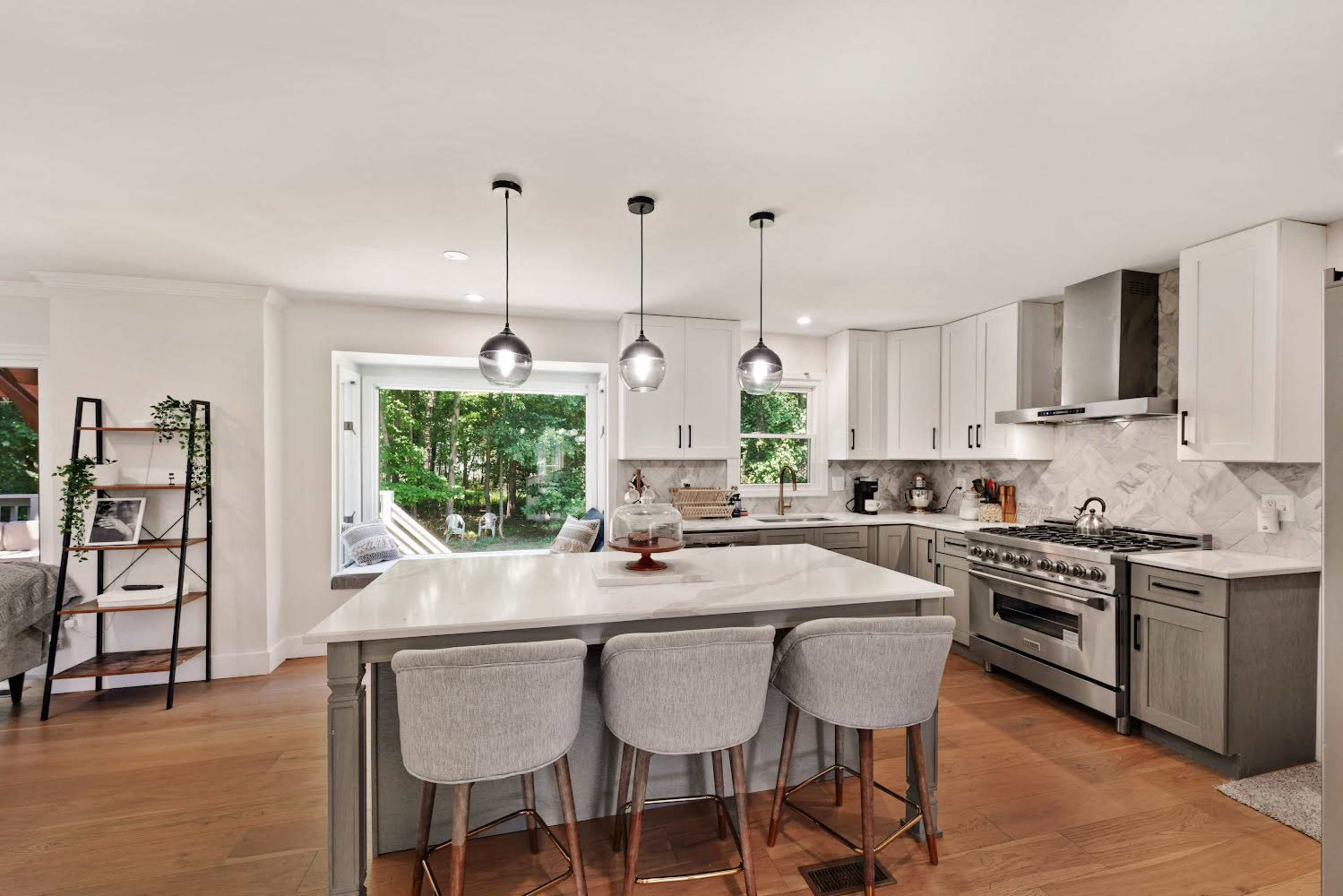 A modern kitchen features a large island with bar seating, sleek cabinetry, and large windows overlooking a wooded area.