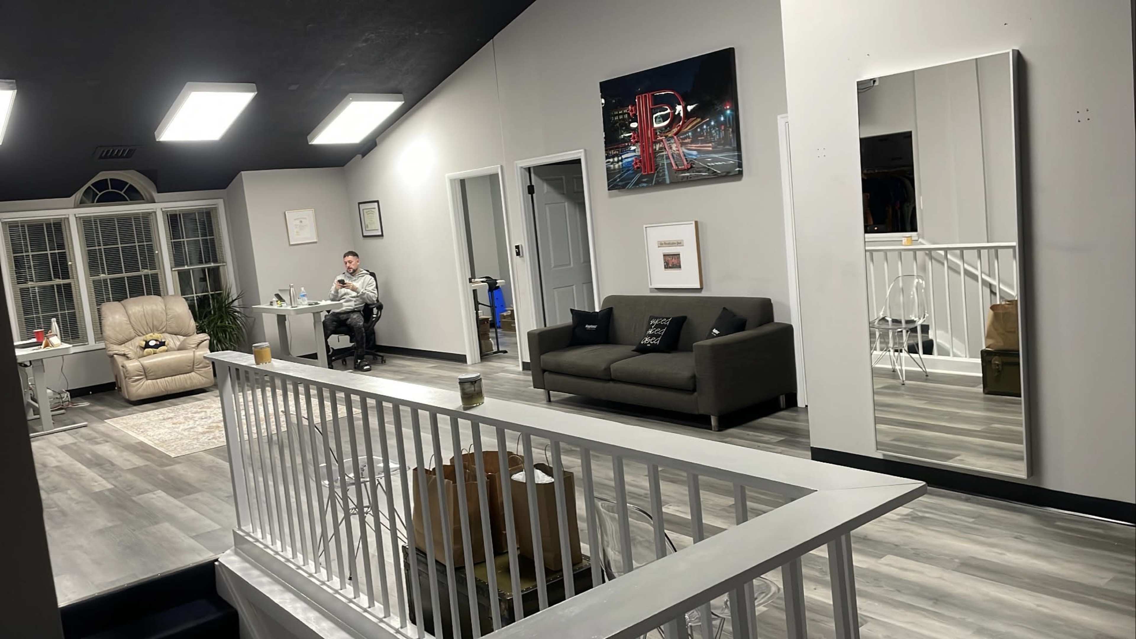 A spacious room features a desk with a person working, a gray sofa, a large mirror, and artwork on the walls.