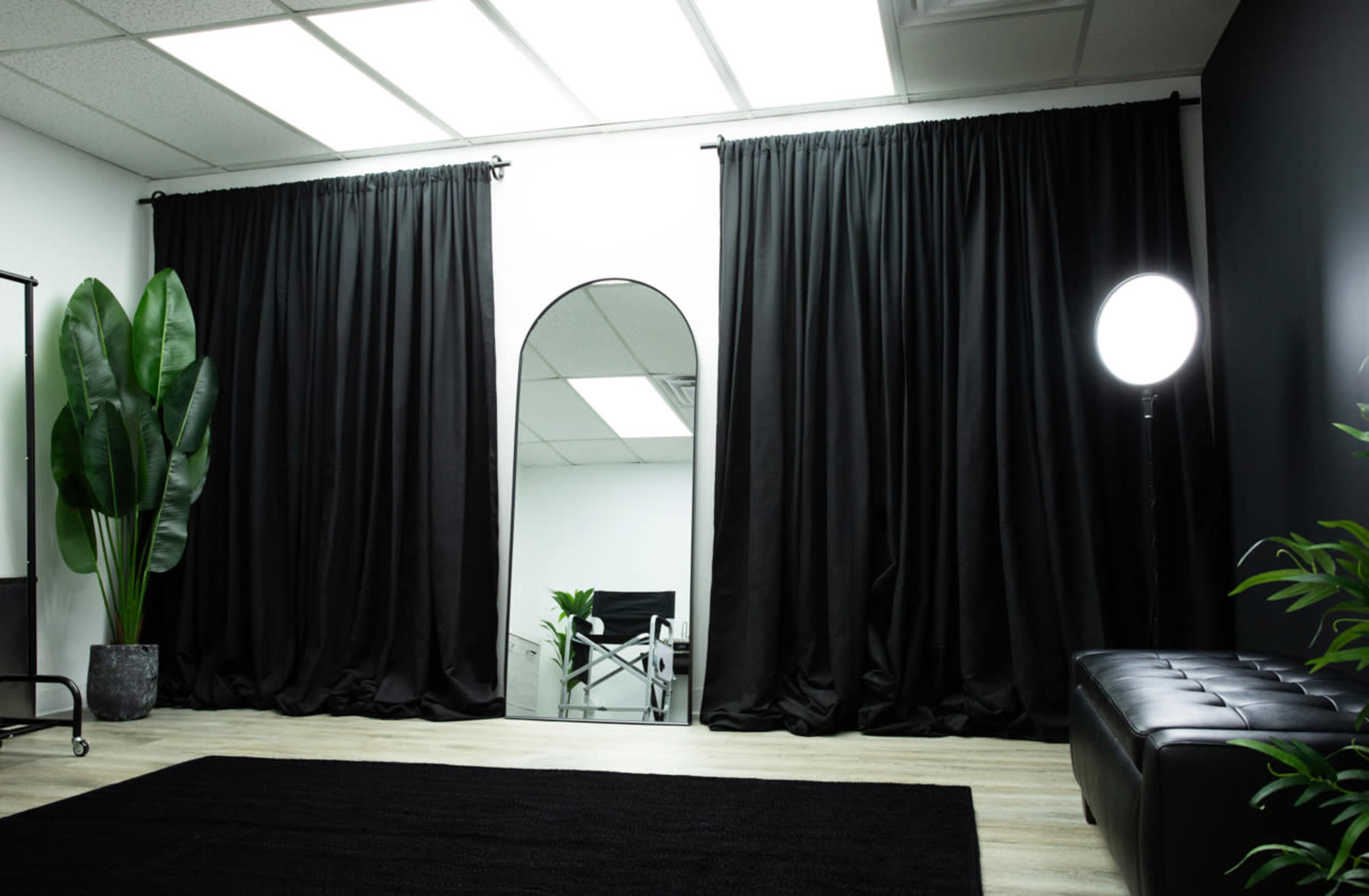 The image shows a sleek interior space with black curtains, a mirror, a large light source, decorative plants, and a dark rug.