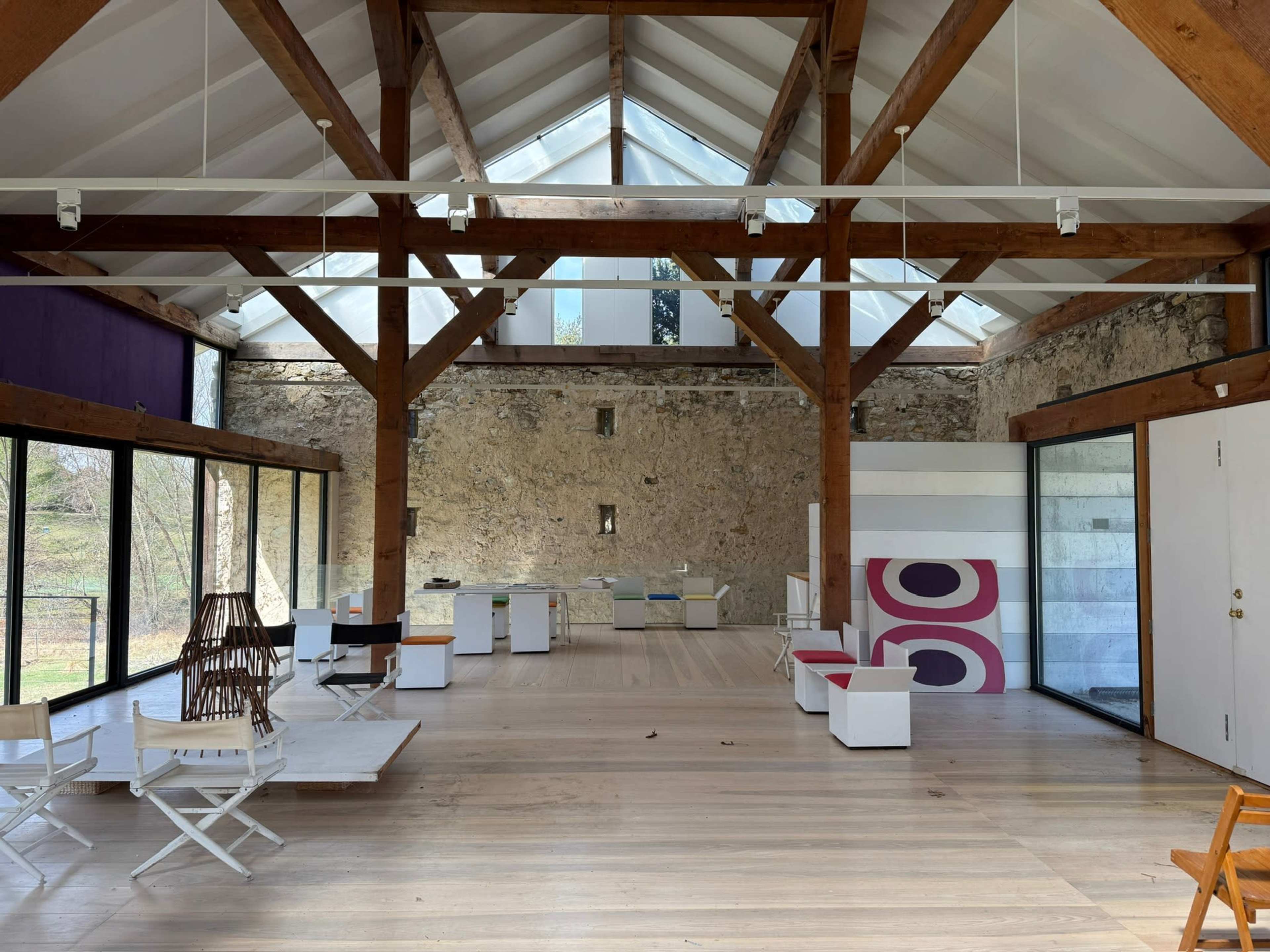 The image shows a spacious interior of a modern building with wooden beams, large windows, and minimalistic furniture, featuring an open layout and a stone wall.