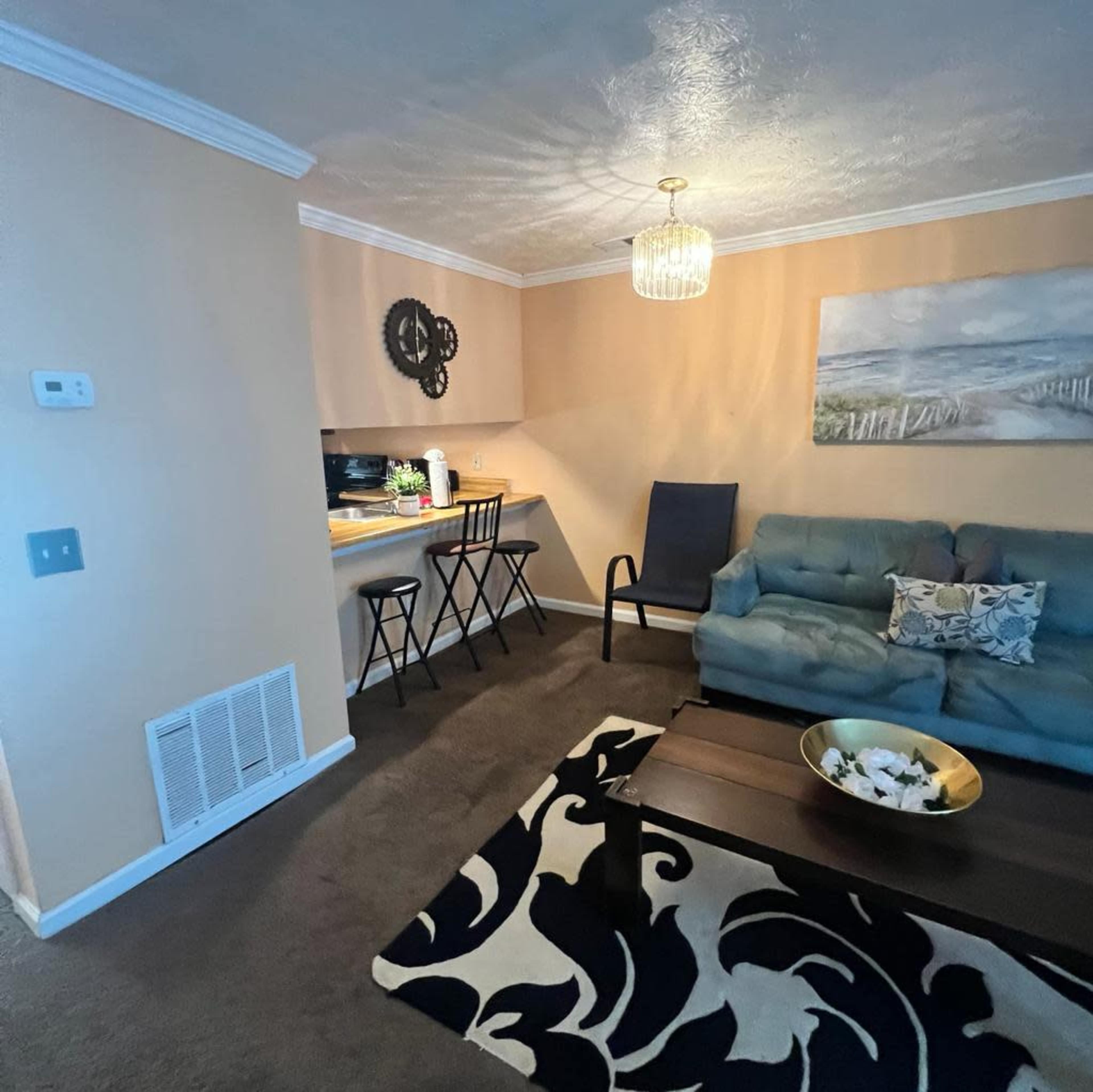 A living area featuring a blue sofa, a black coffee table on a patterned rug, and a small dining setup with two stools near a kitchen area.