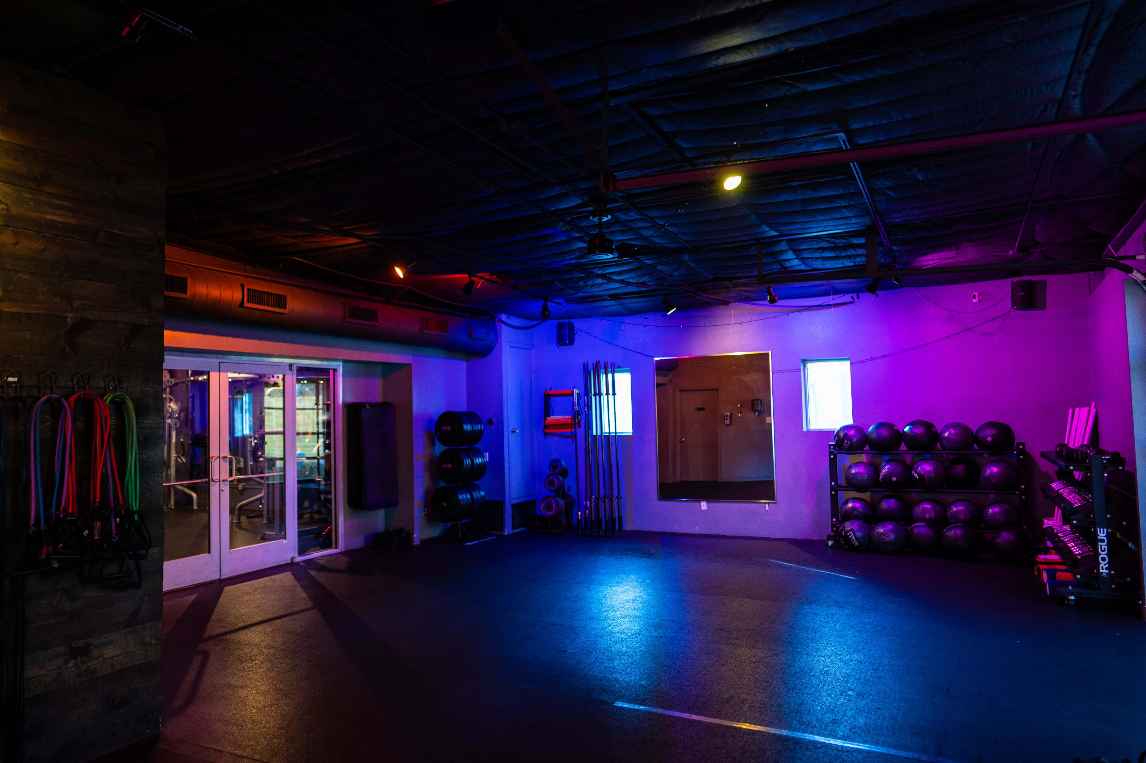 A dimly lit gym interior with colorful lighting, featuring exercise equipment along the walls and free weights stacked in one corner.