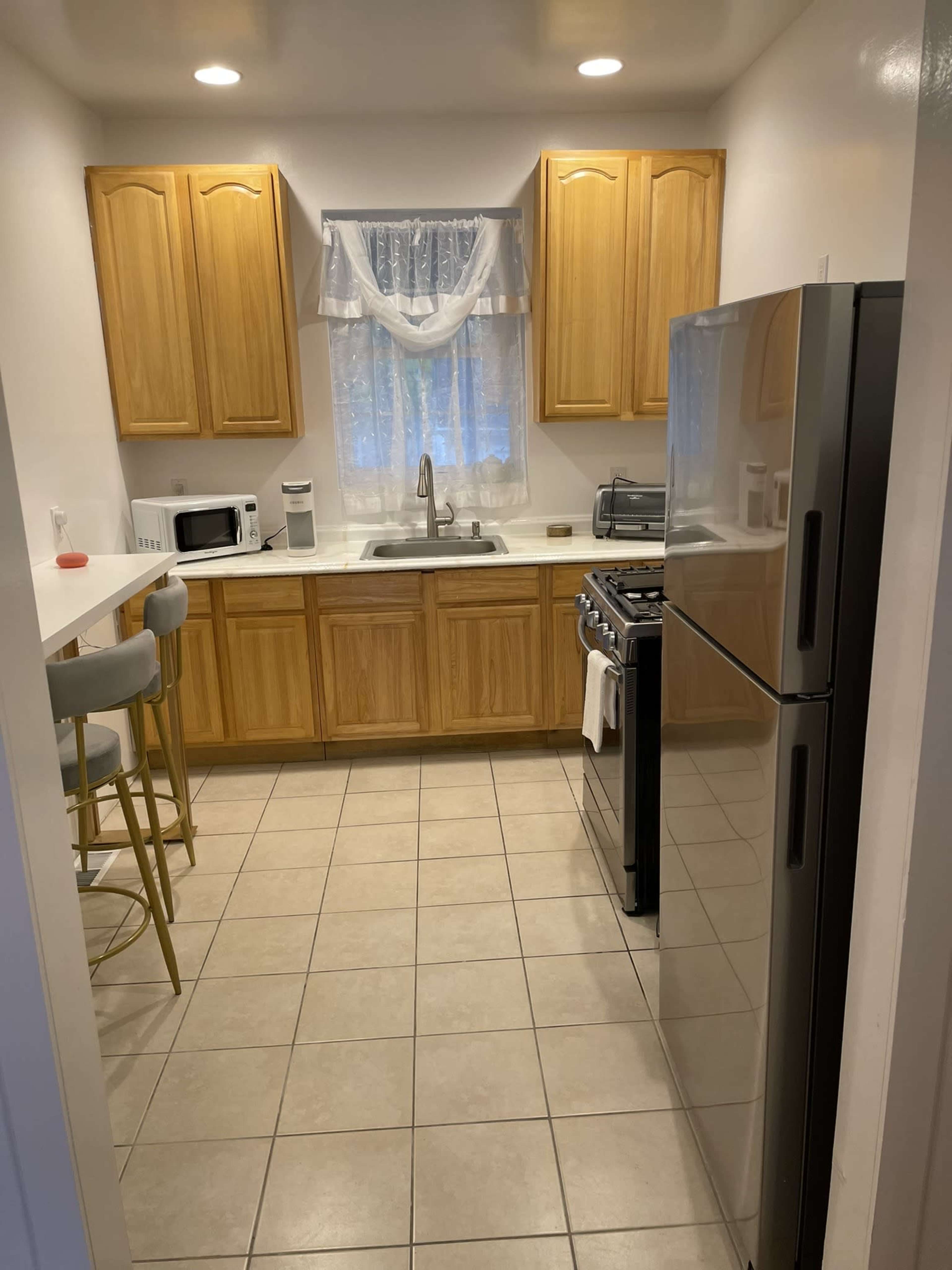 A kitchen with wooden cabinets, a sink under a window, a gas stove, a refrigerator, and a small breakfast bar with three stools.