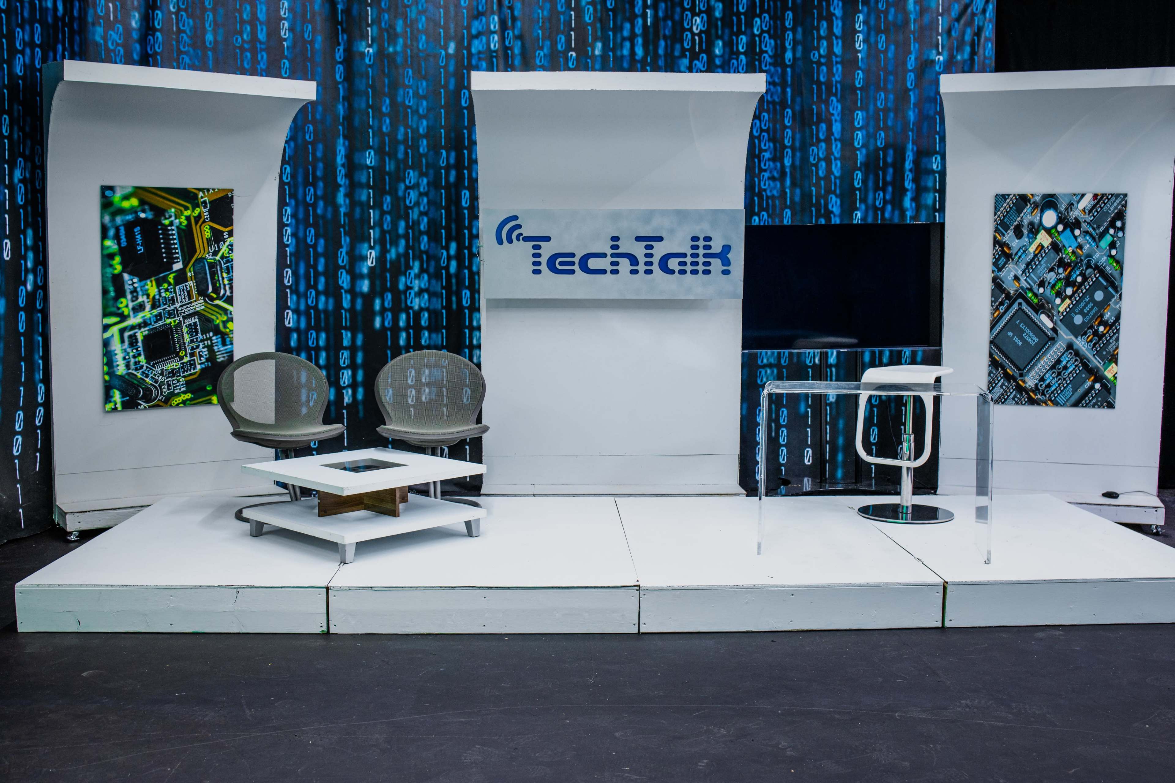 The image shows a modern television studio setup for a tech-themed program, featuring two chairs, a glass table, and digital-themed backdrops with circuit board designs.