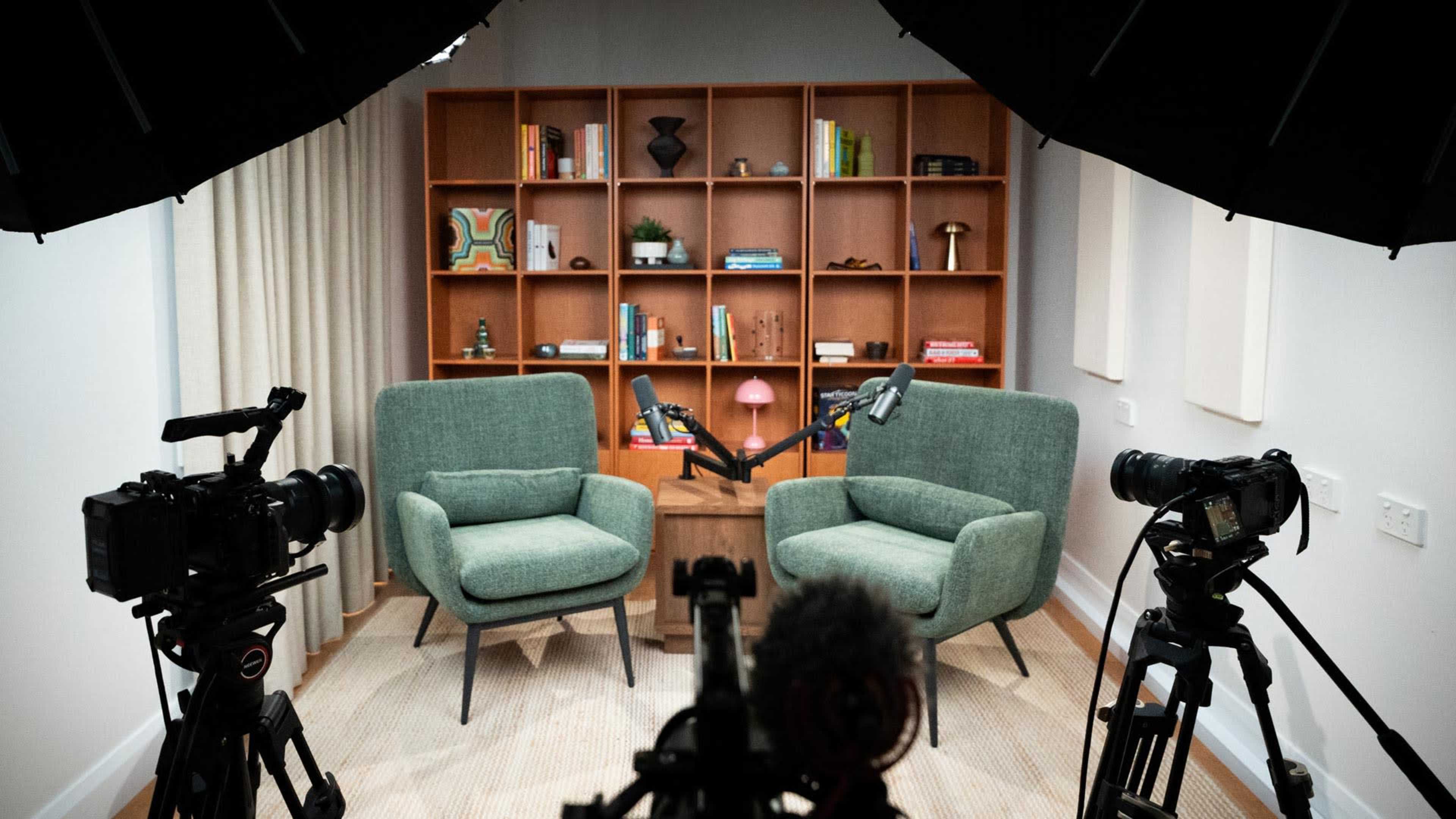 The image shows a well-lit podcast studio setup featuring two green armchairs facing each other, with a wooden table in between and cameras positioned on either side.