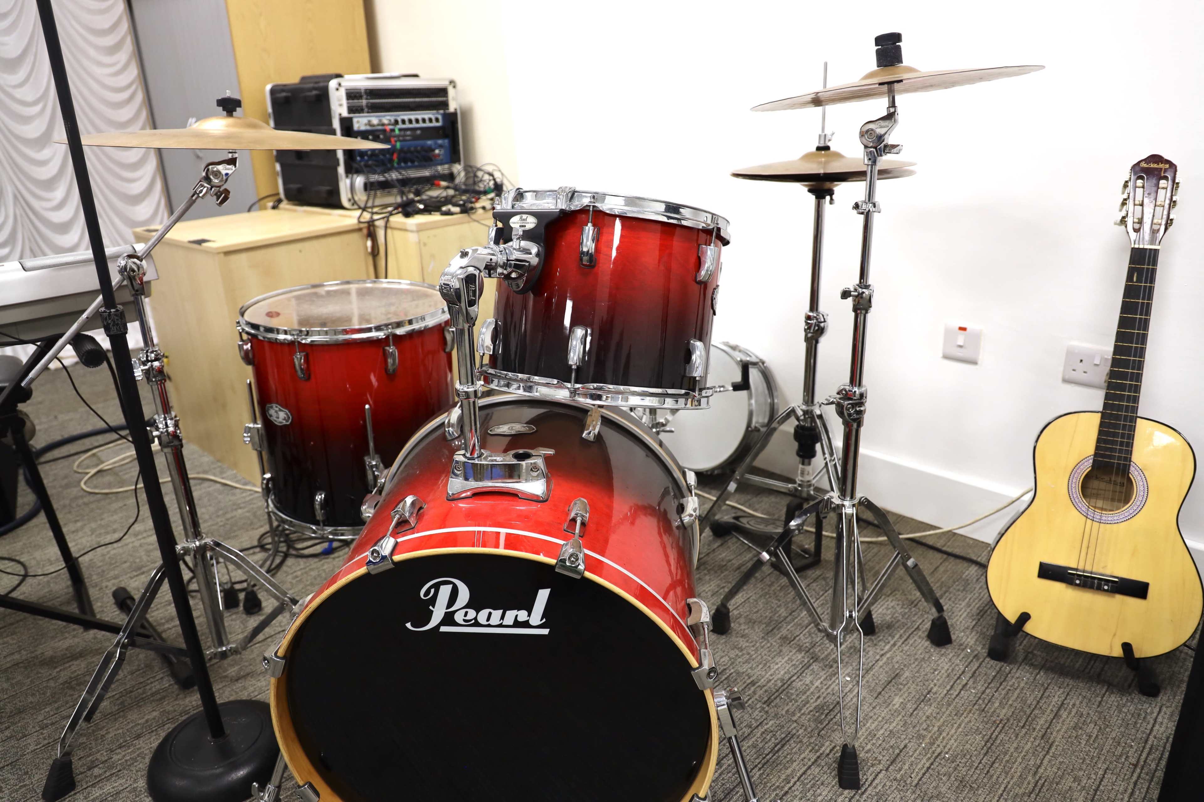The image shows a red Pearl drum kit, including a bass drum and a snare drum, alongside a guitar and audio equipment in a well-lit room.