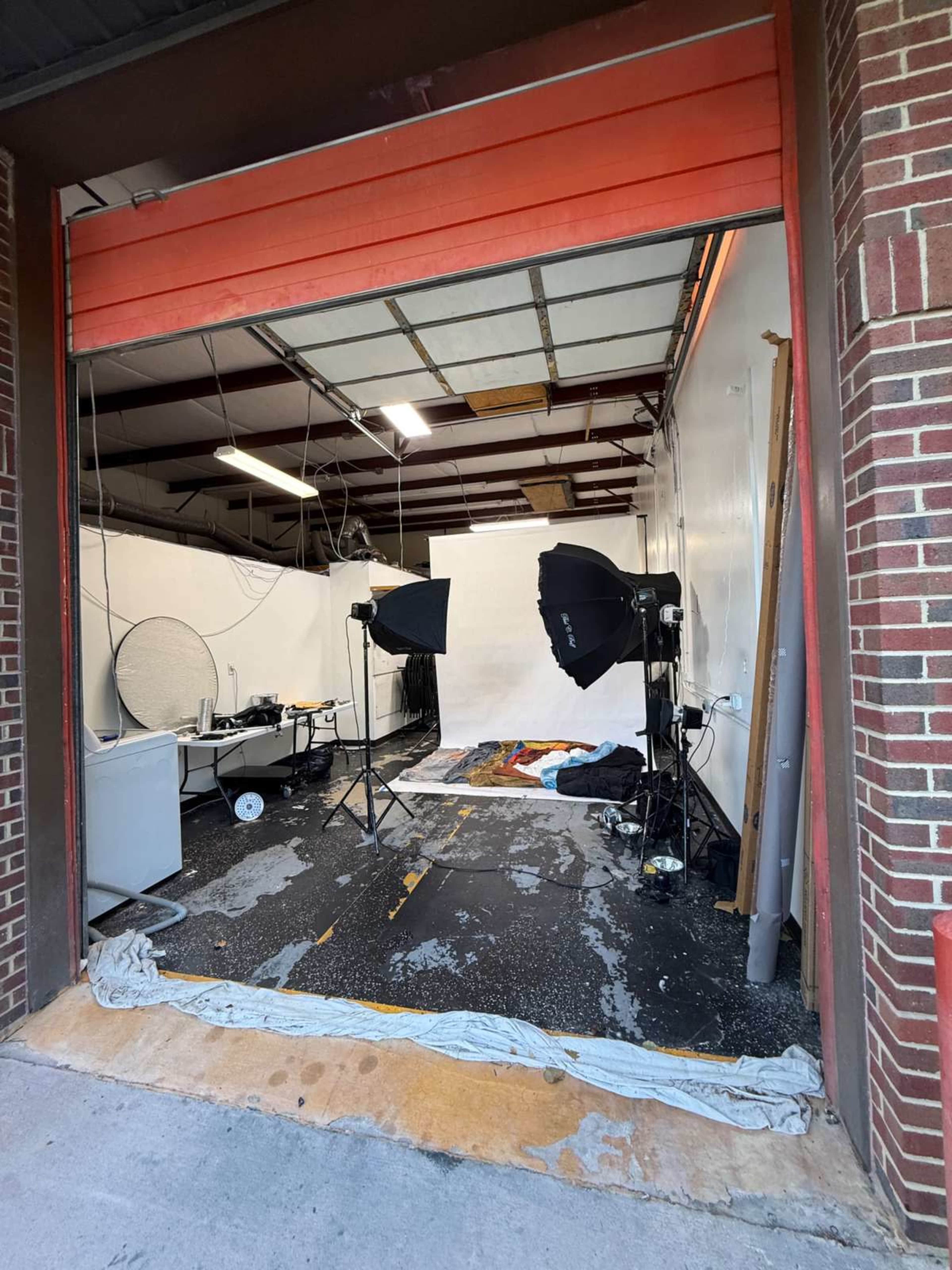 The image shows an open garage space set up as a photography studio, featuring backdrop equipment, lighting, and various props on the floor.