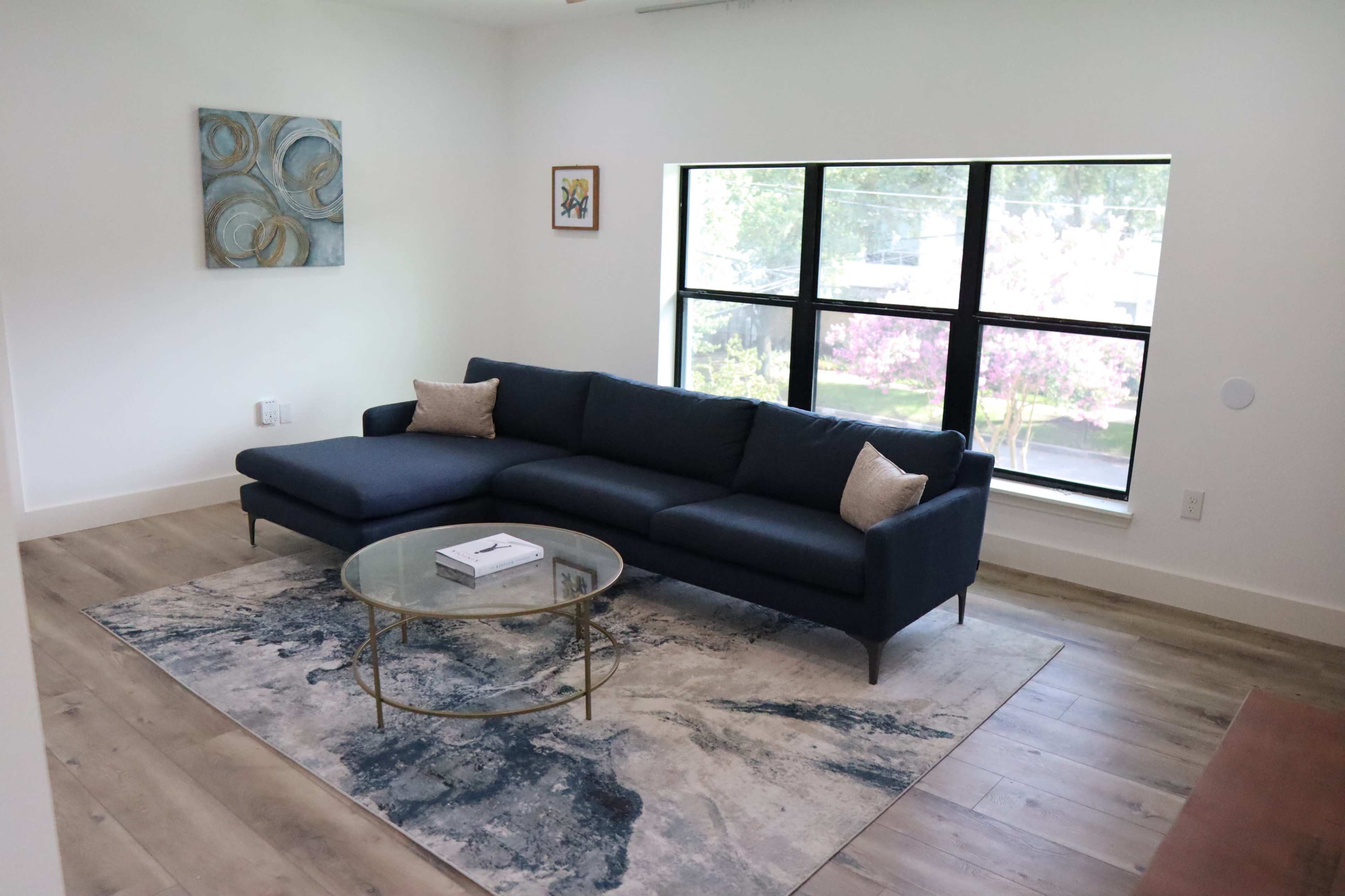 A modern living room featuring a dark blue sectional sofa, a glass coffee table, and a large window with a view of blooming trees outside.