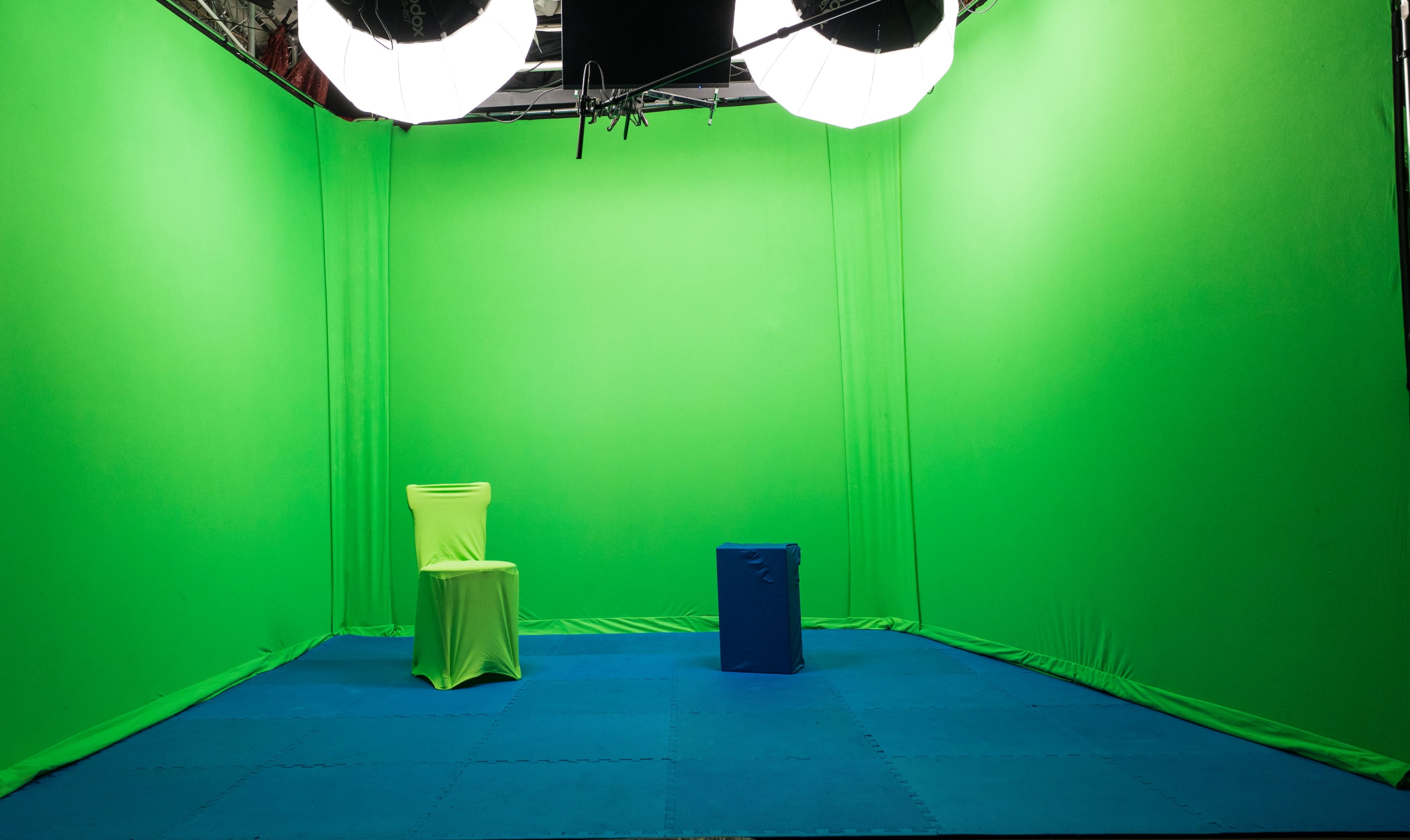 The image shows an empty studio space with a green backdrop, two lights hanging from the ceiling, and a blue mat on the floor.