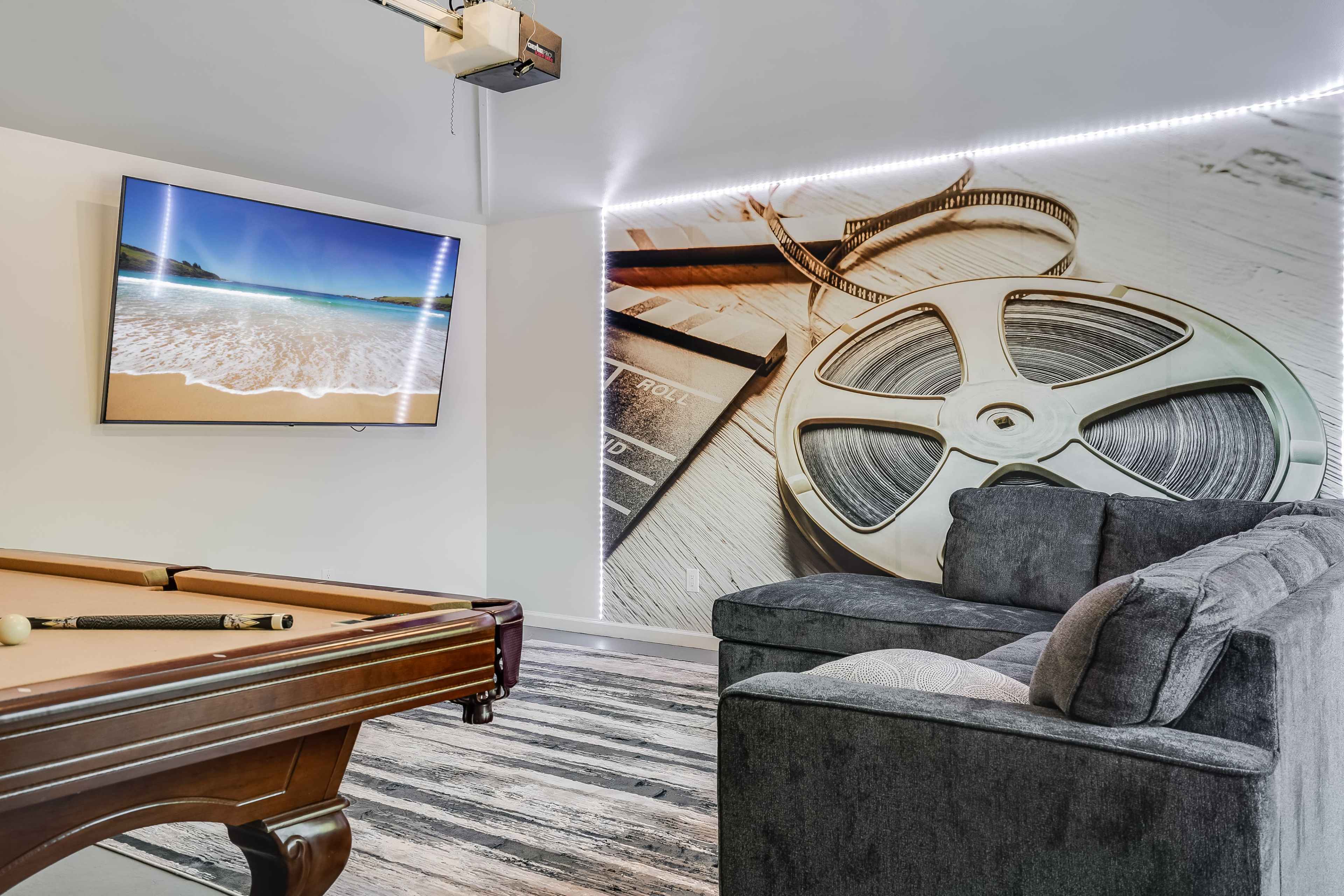 A game room features a billiards table, a television displaying a beach scene, and a textured wall with film reel imagery.