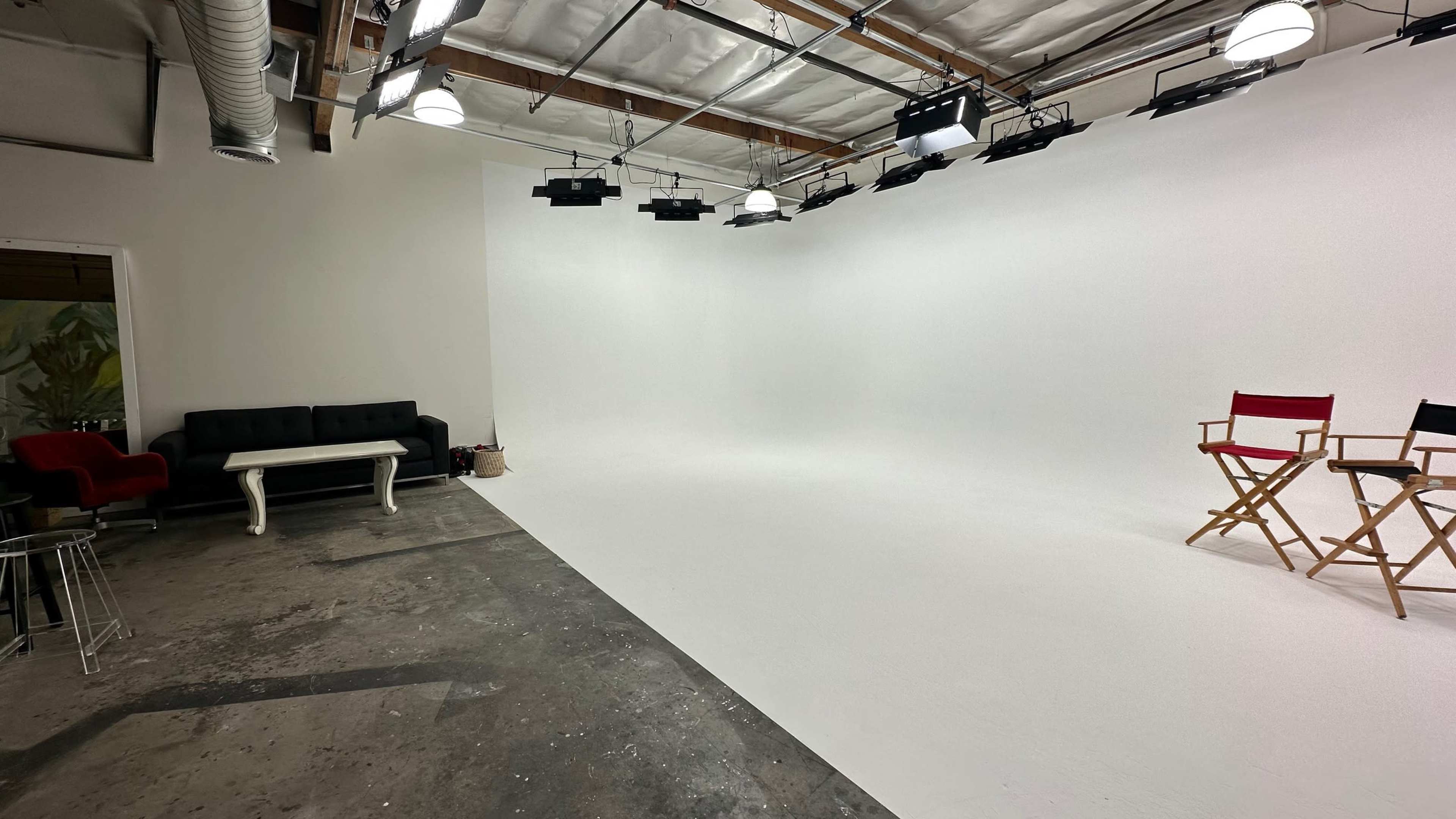 A spacious studio with a white backdrop, two director's chairs, a black sofa, and overhead lighting.