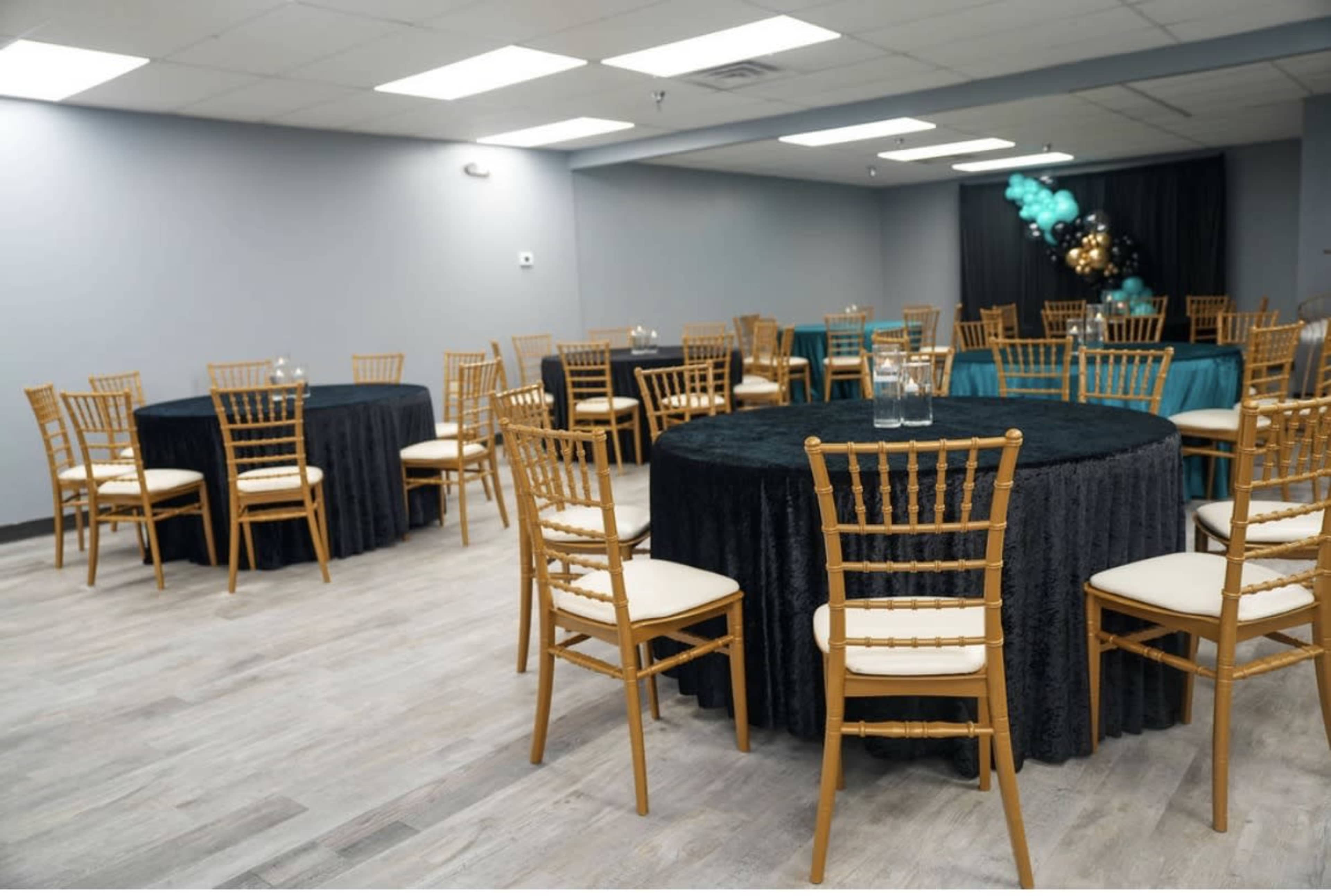 The image shows a banquet hall with several round tables covered in black and teal tablecloths, surrounded by wooden chairs.