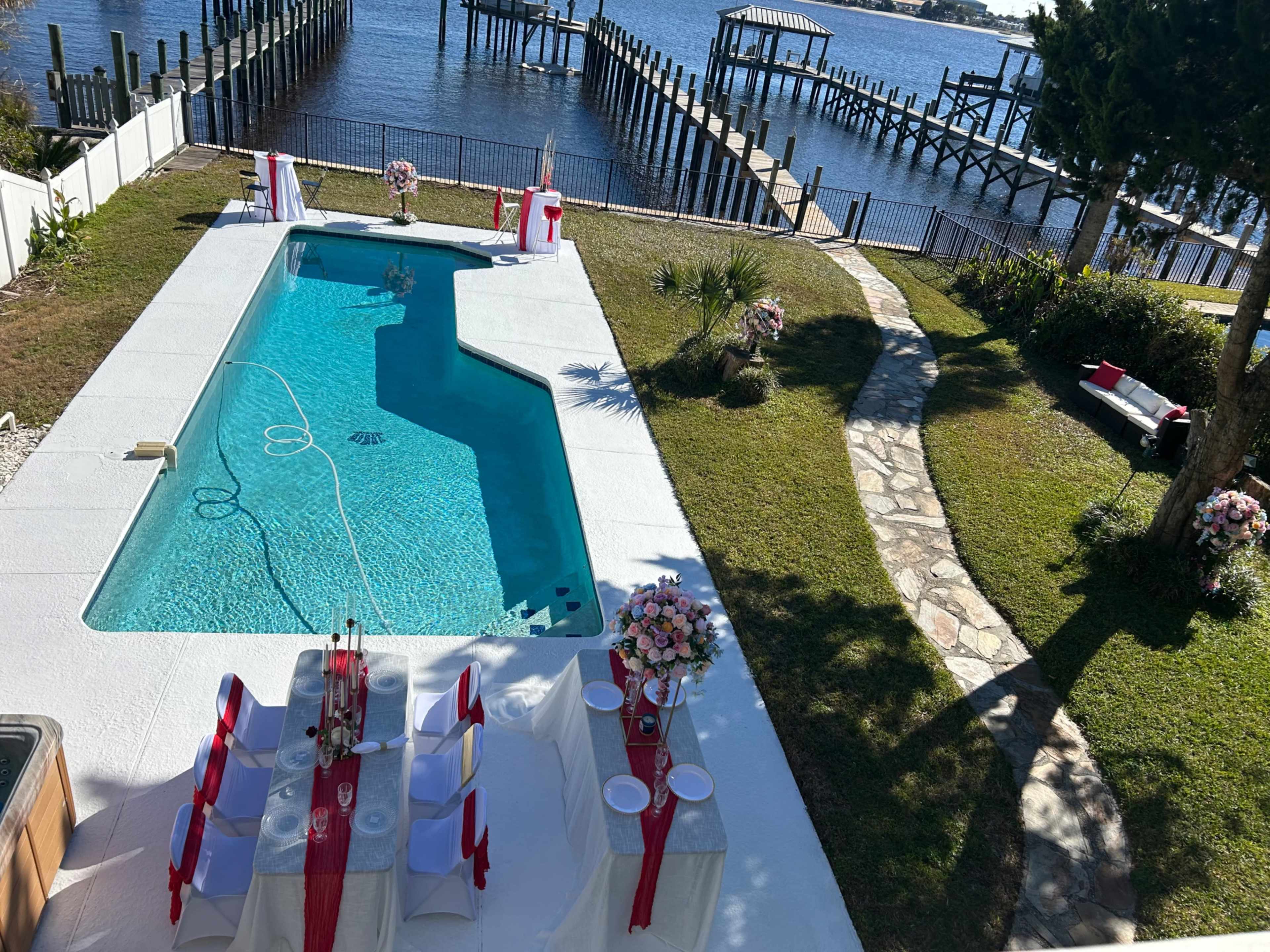 The image shows a backyard pool area with a table set for an event, overlooking a body of water and a dock in the background.