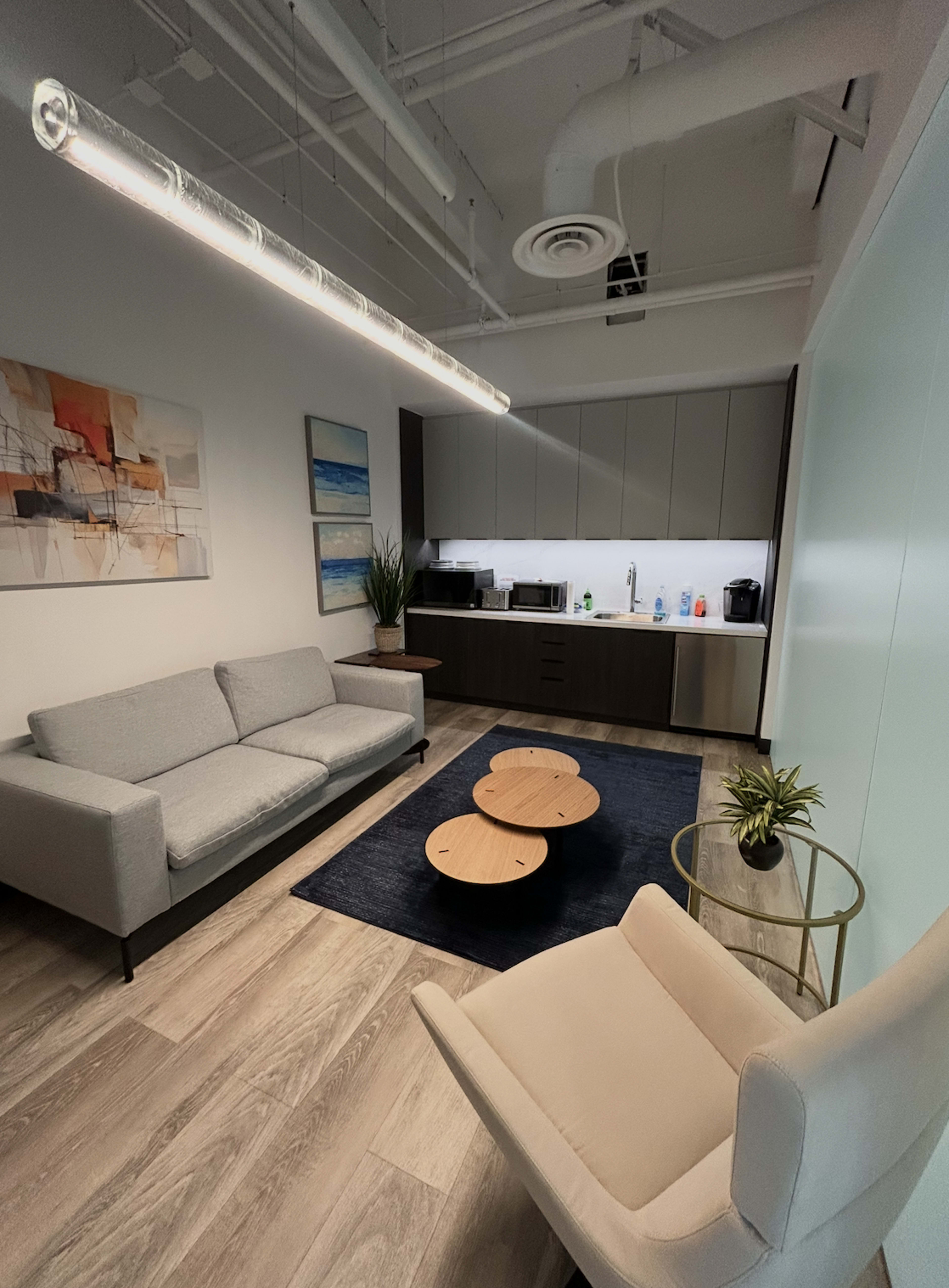 The image shows a modern office break room with a gray couch, a round coffee table, a light-colored armchair, and a kitchenette in the background.