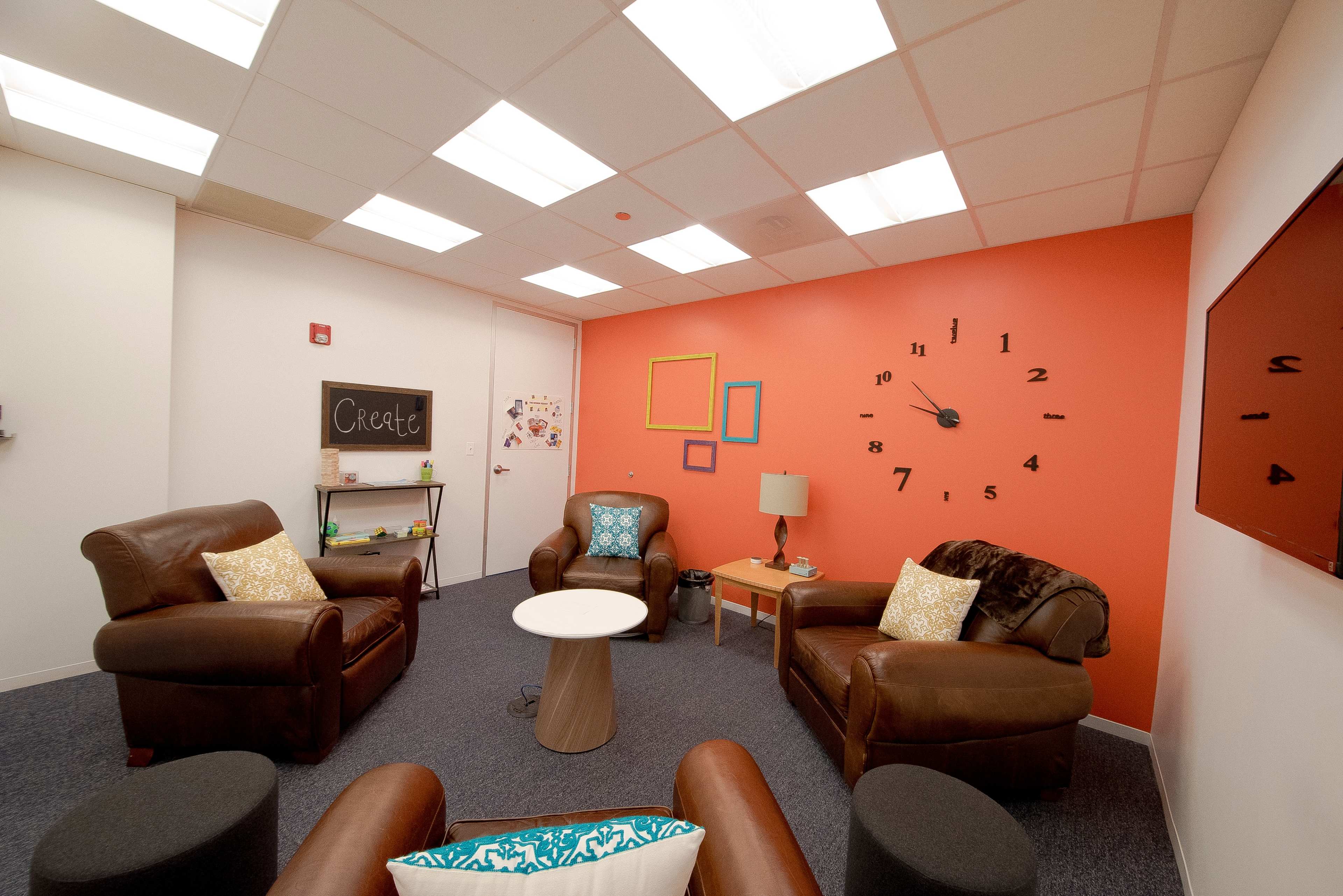 The image shows a cozy meeting room with leather chairs, a round table, a wall clock, and colorful framed decorations.