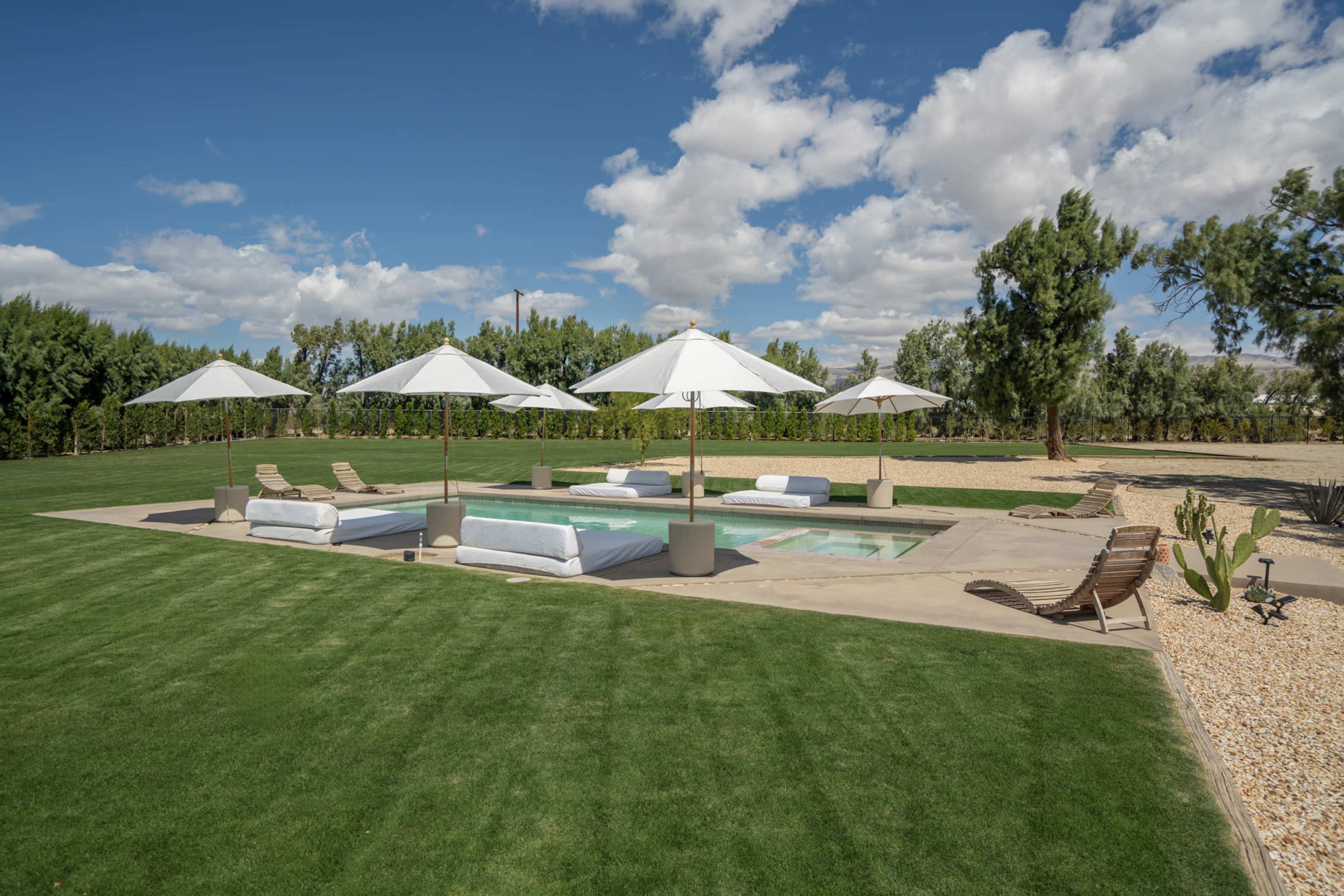 A landscaped outdoor area features a rectangular pool surrounded by lounge chairs and umbrellas on a green lawn under a partly cloudy sky.