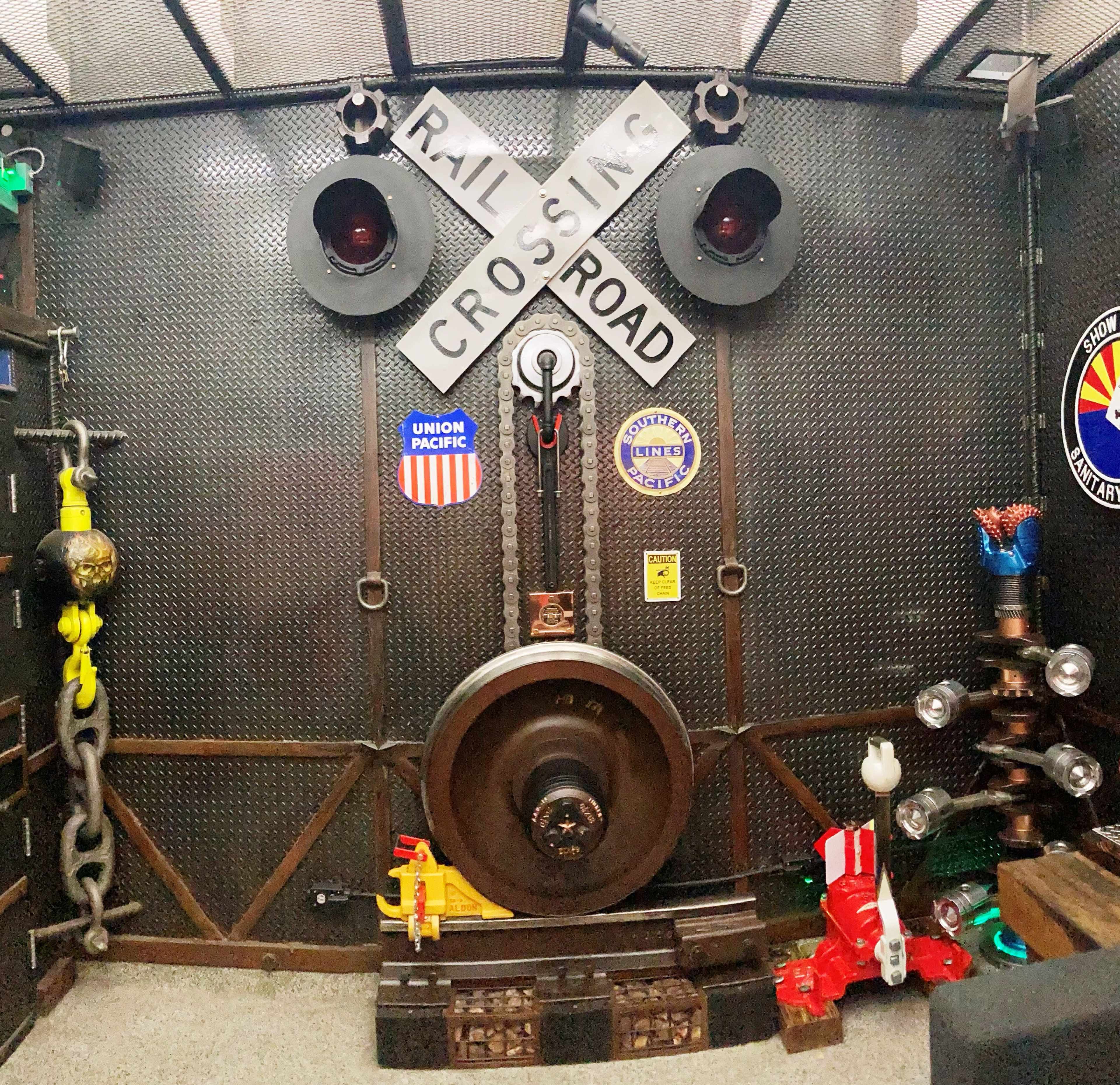 The image shows a wall featuring a railroad crossing sign, traffic lights, and various railroad-themed decorations, including chains and a large wheel.