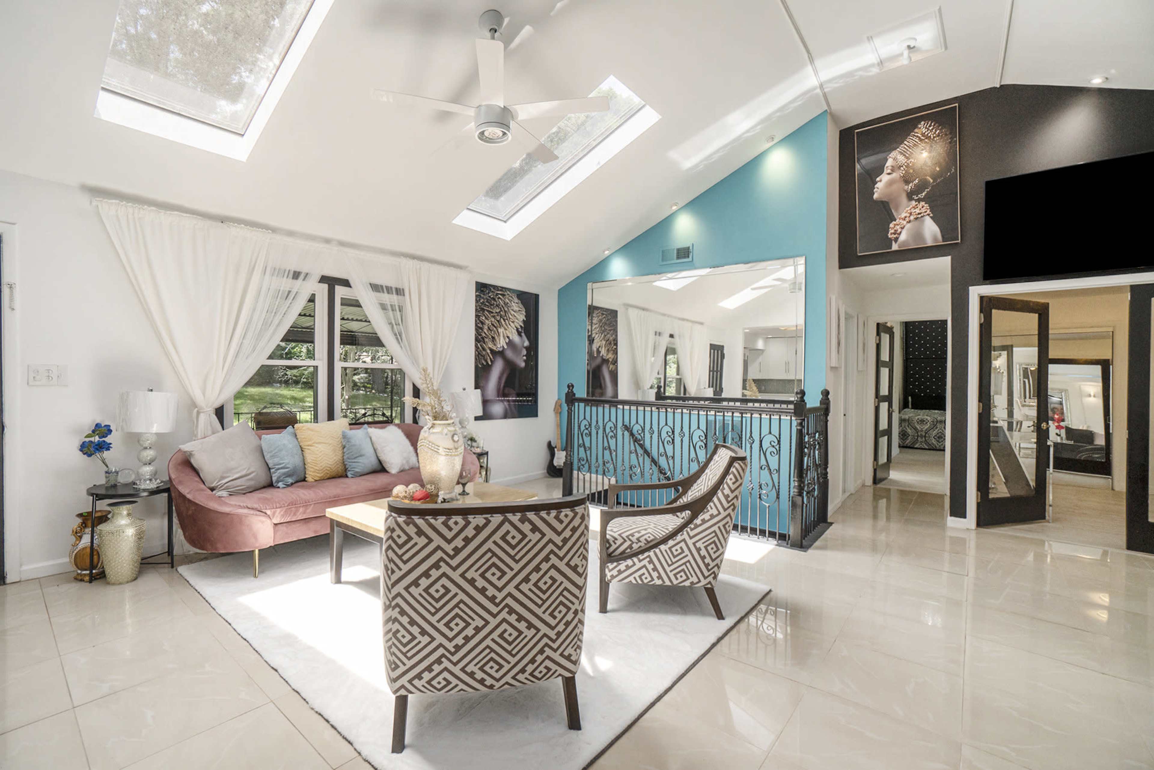 A bright, modern living room featuring a pink sofa and patterned armchairs, with large skylights and decorative wall art.