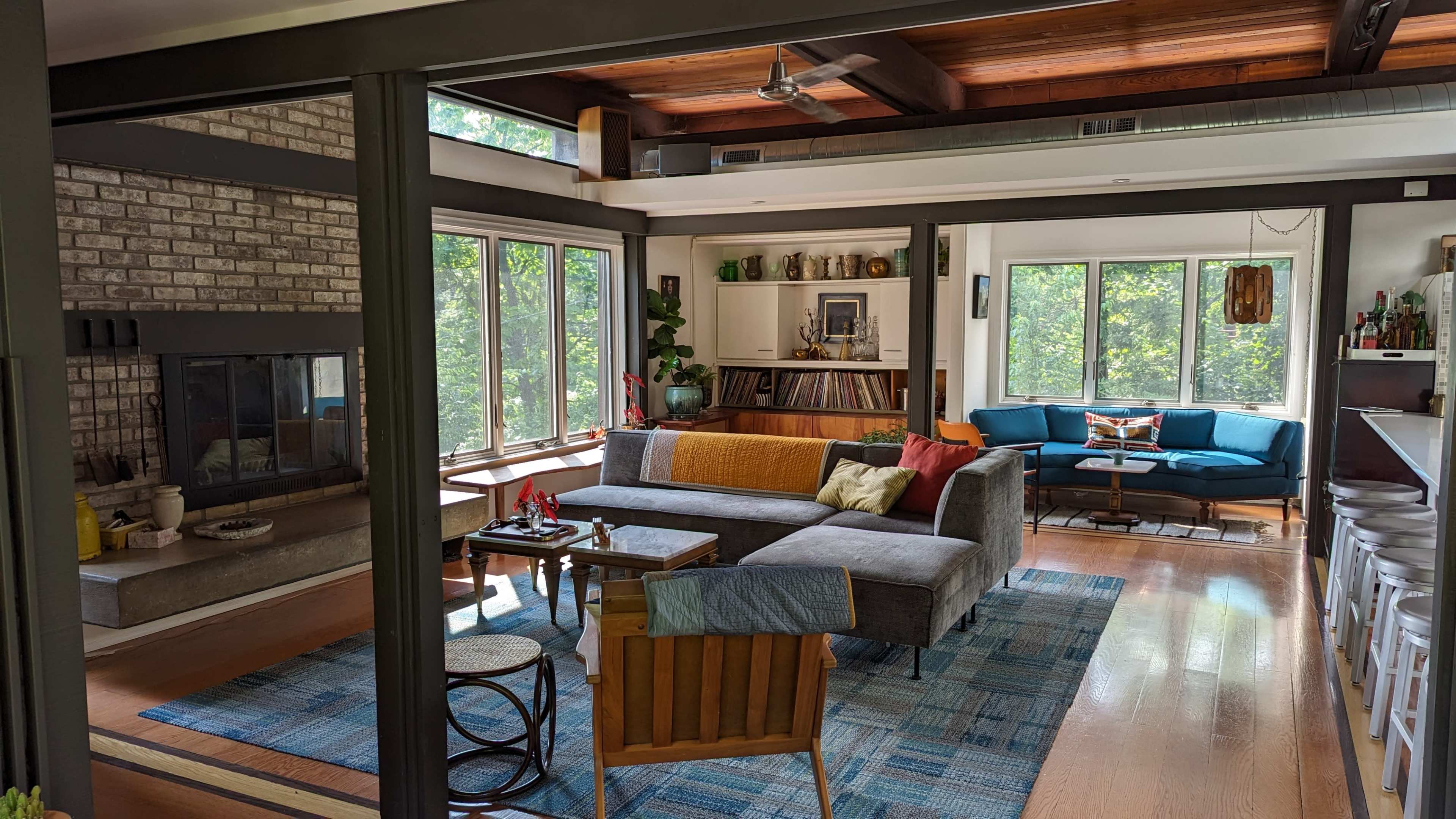 The image shows a modern living room featuring a gray sectional sofa, a brick fireplace, large windows, and a mix of wooden and upholstered furniture.