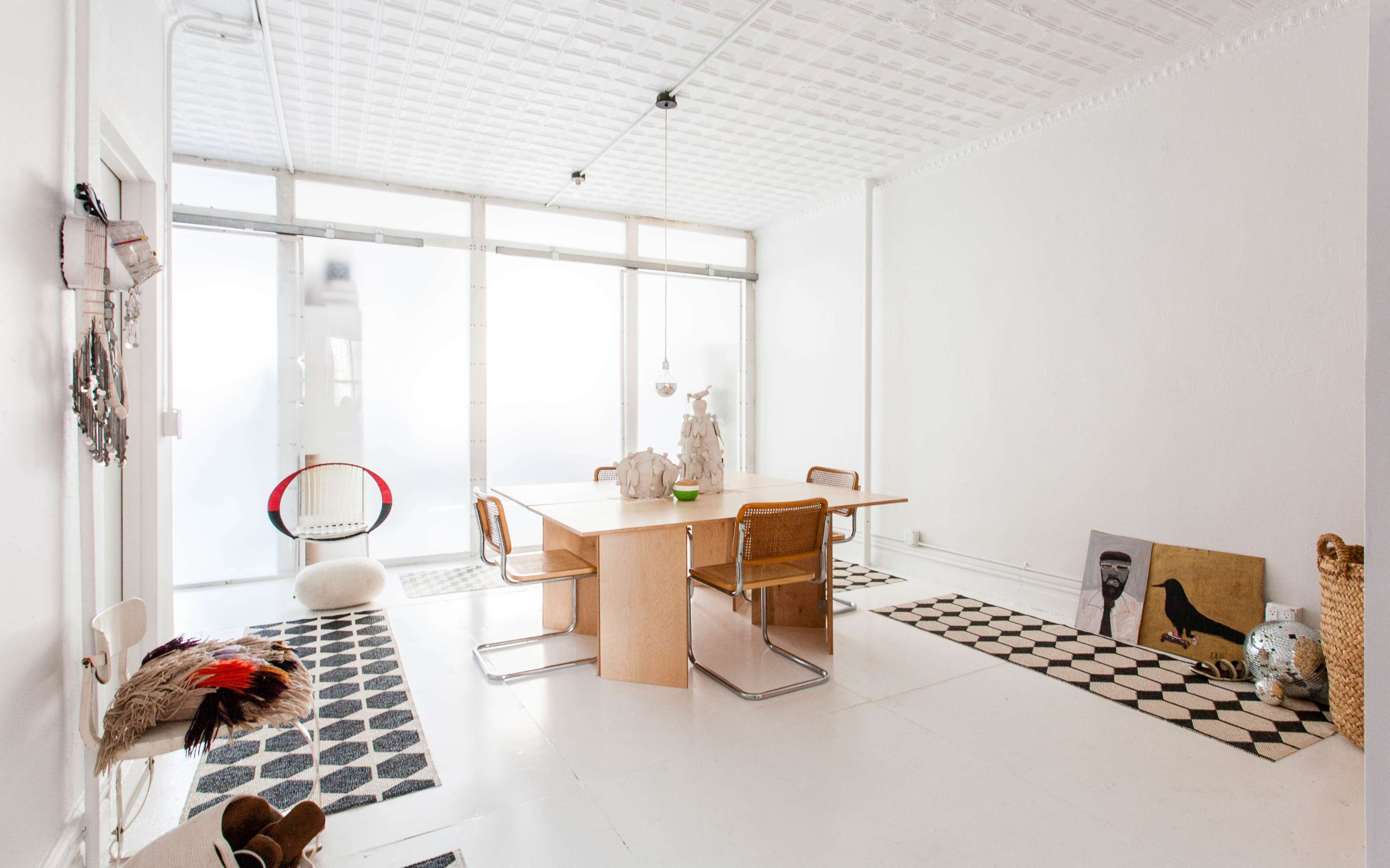 A minimalistic dining area featuring a wooden table surrounded by chairs, with decorative rugs on the floor and large windows allowing light to filter in.