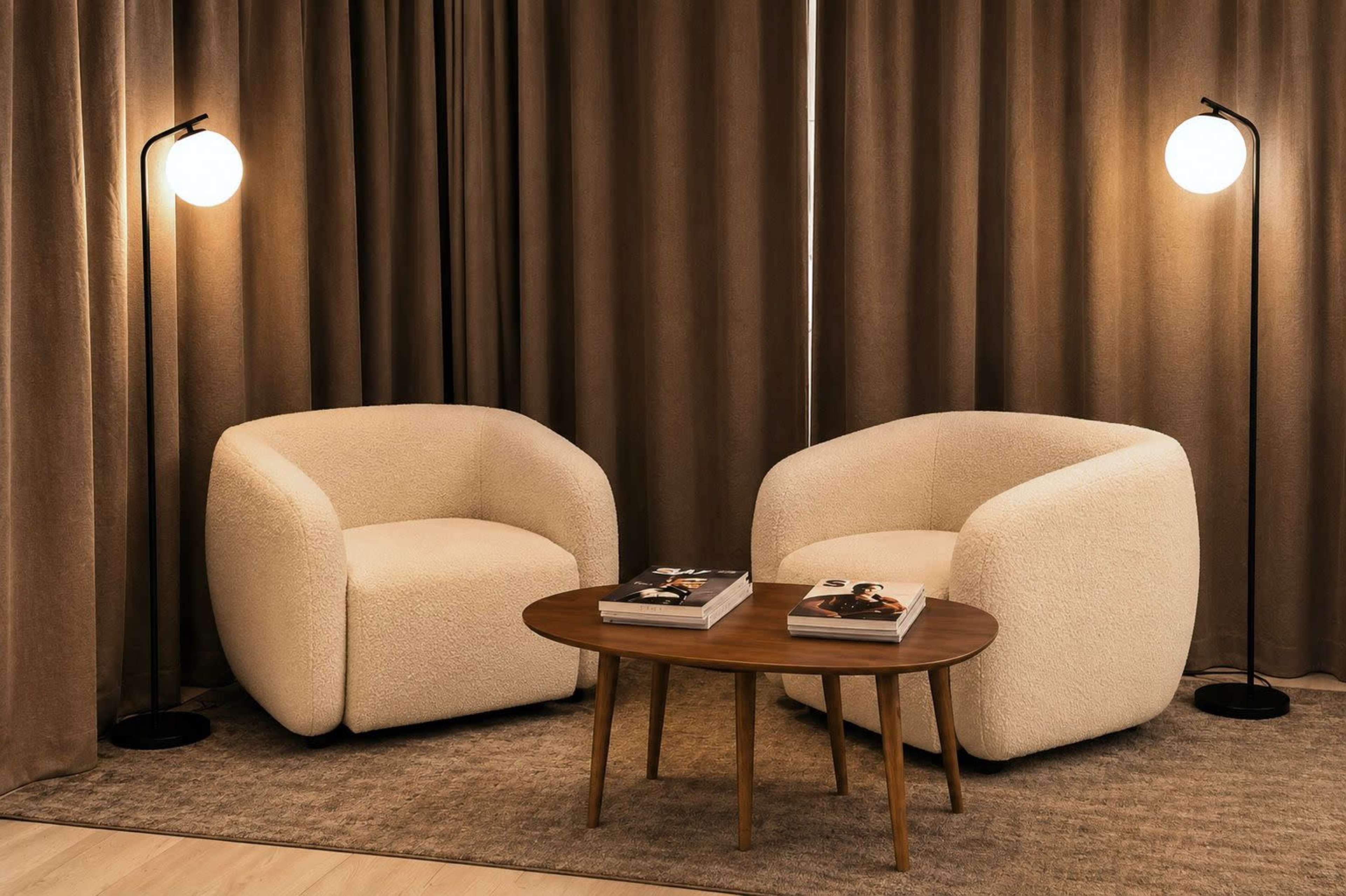 The image shows two round, white upholstered chairs flanking a wooden coffee table, with two floor lamps providing light against a backdrop of brown curtains.