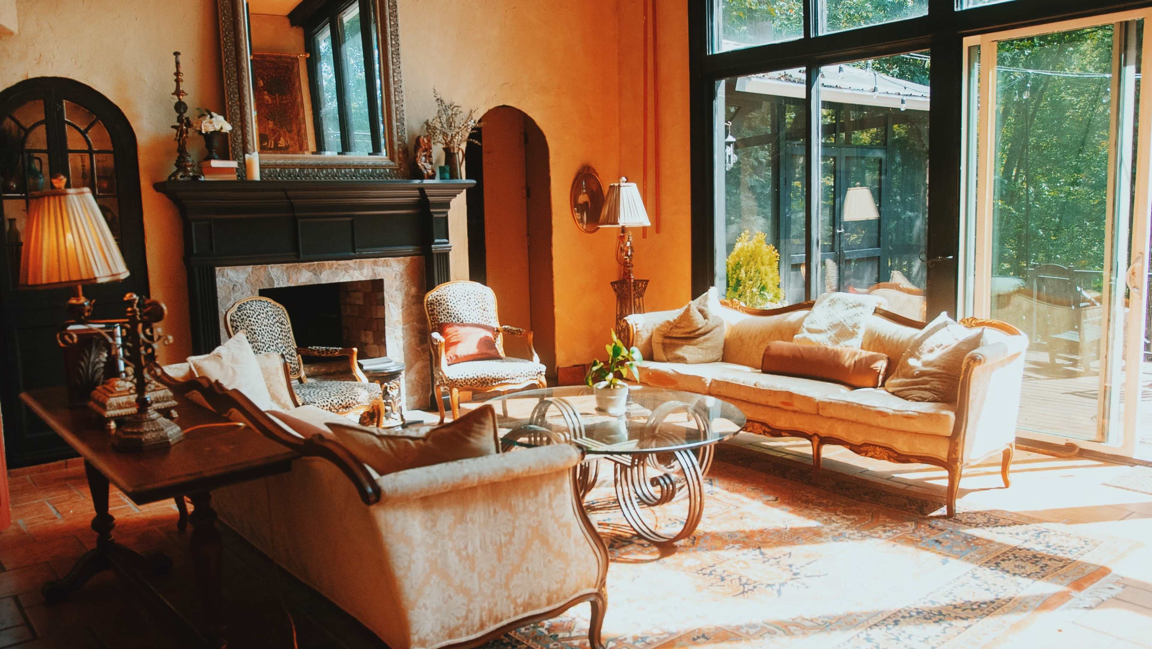 The image shows a cozy living room with a fireplace, two sofas, a coffee table, and large windows that offer a view of a deck and surrounding greenery.