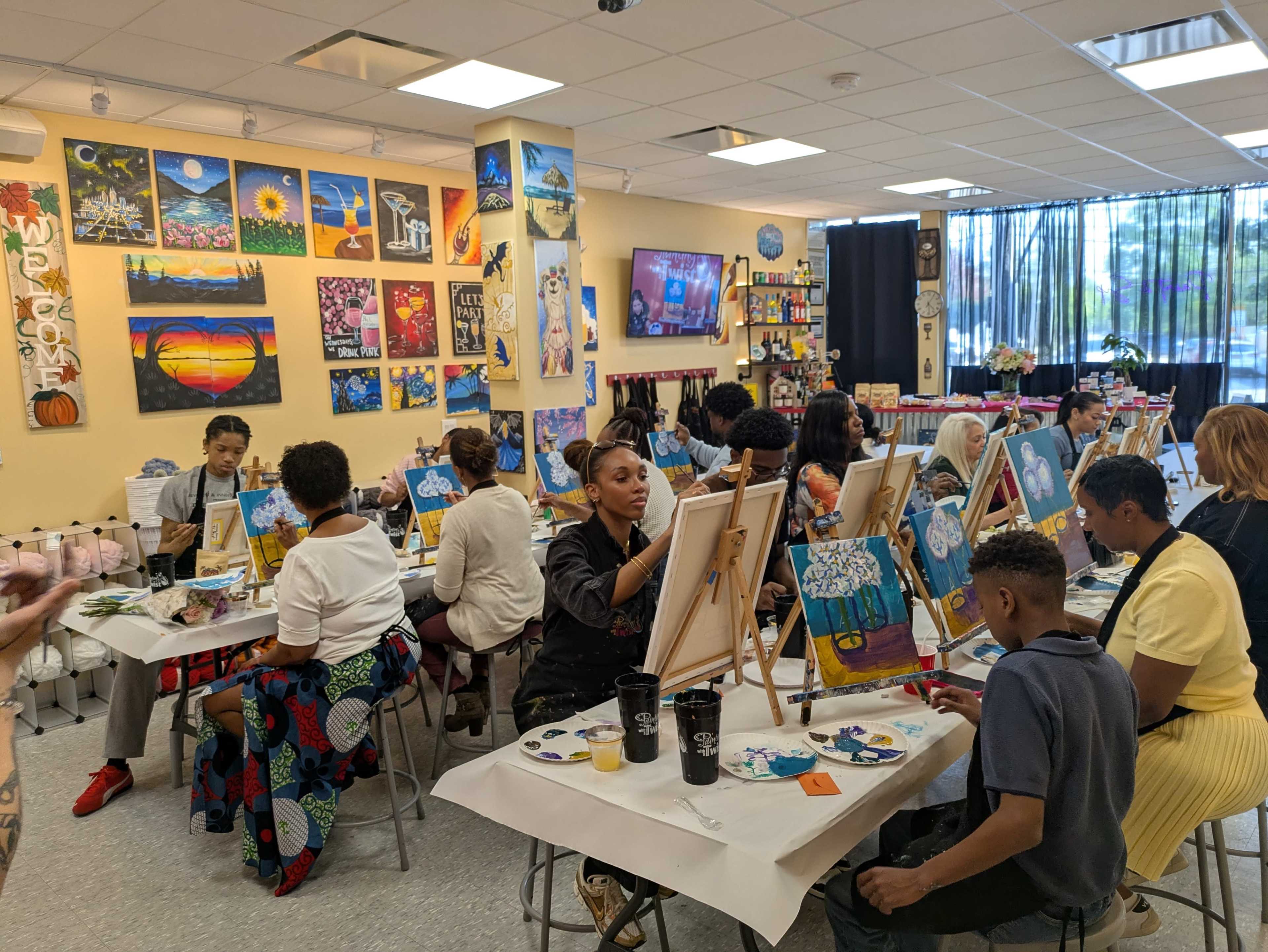 A group of people sits at tables painting on canvases in a brightly decorated art studio.
