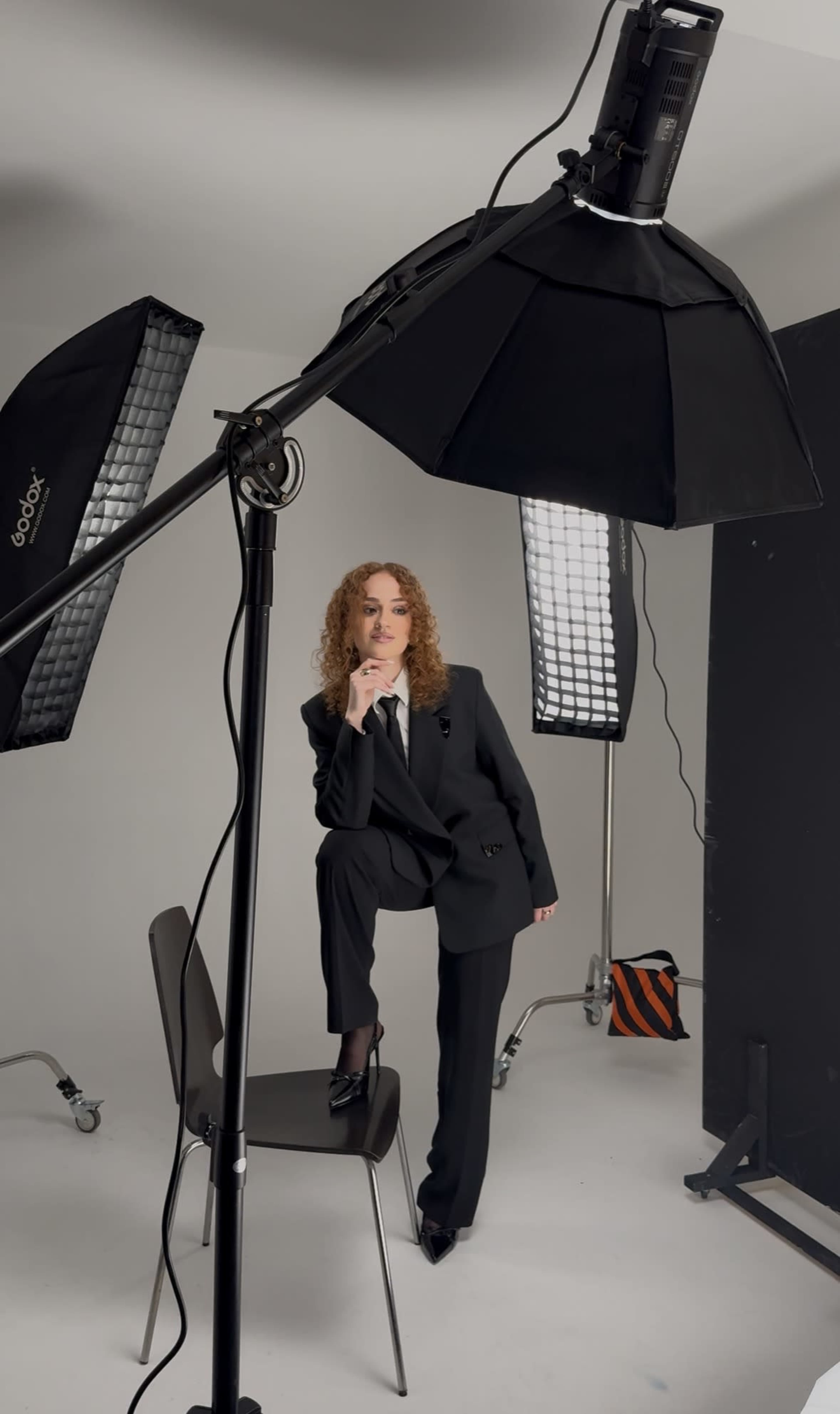 A person wearing a black suit poses on a chair in a photo studio with softbox lights.