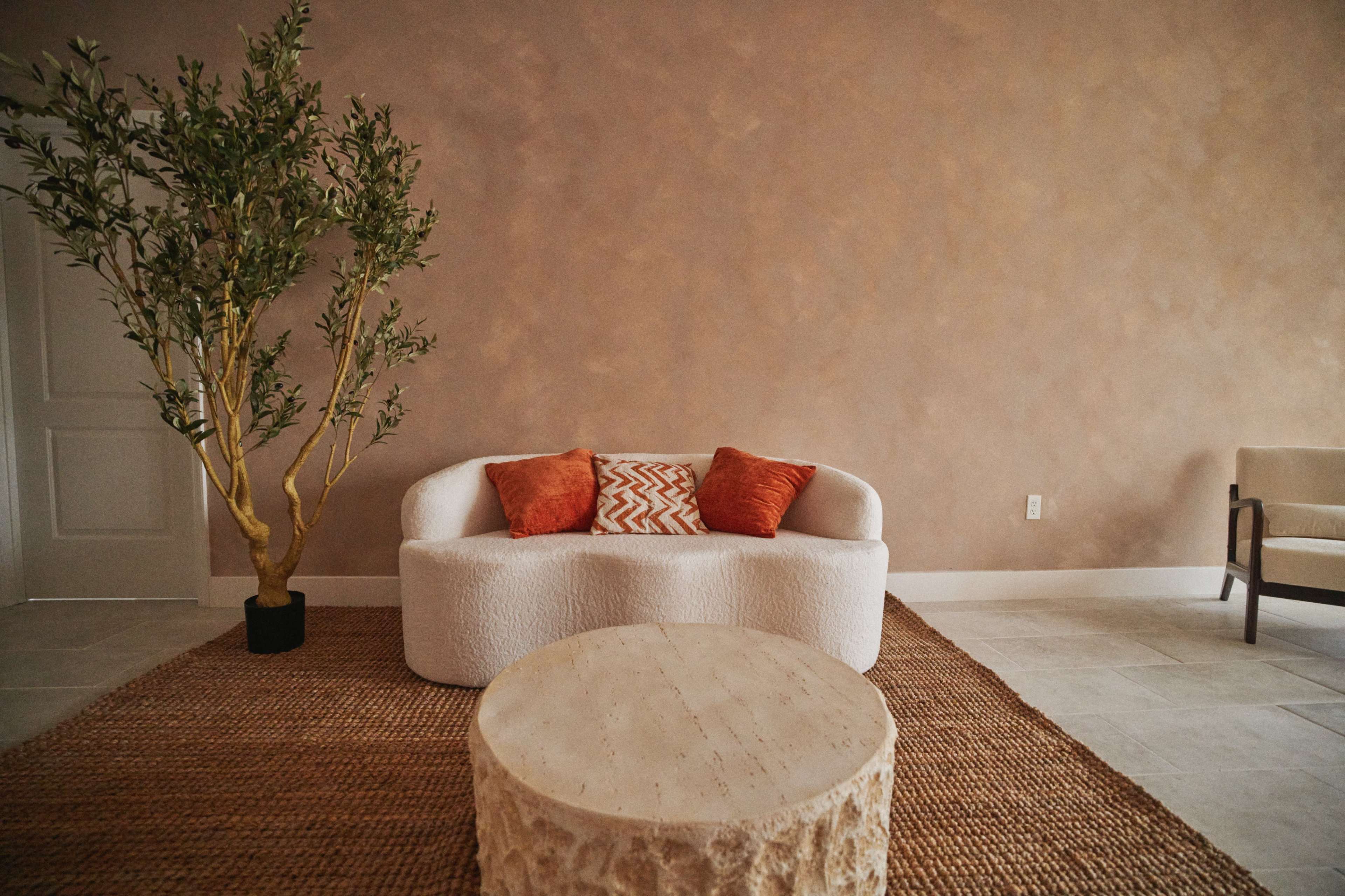 A textured beige wall serves as a backdrop for a white, rounded sofa with orange cushions, complemented by a round wooden coffee table and a potted plant in a corner.