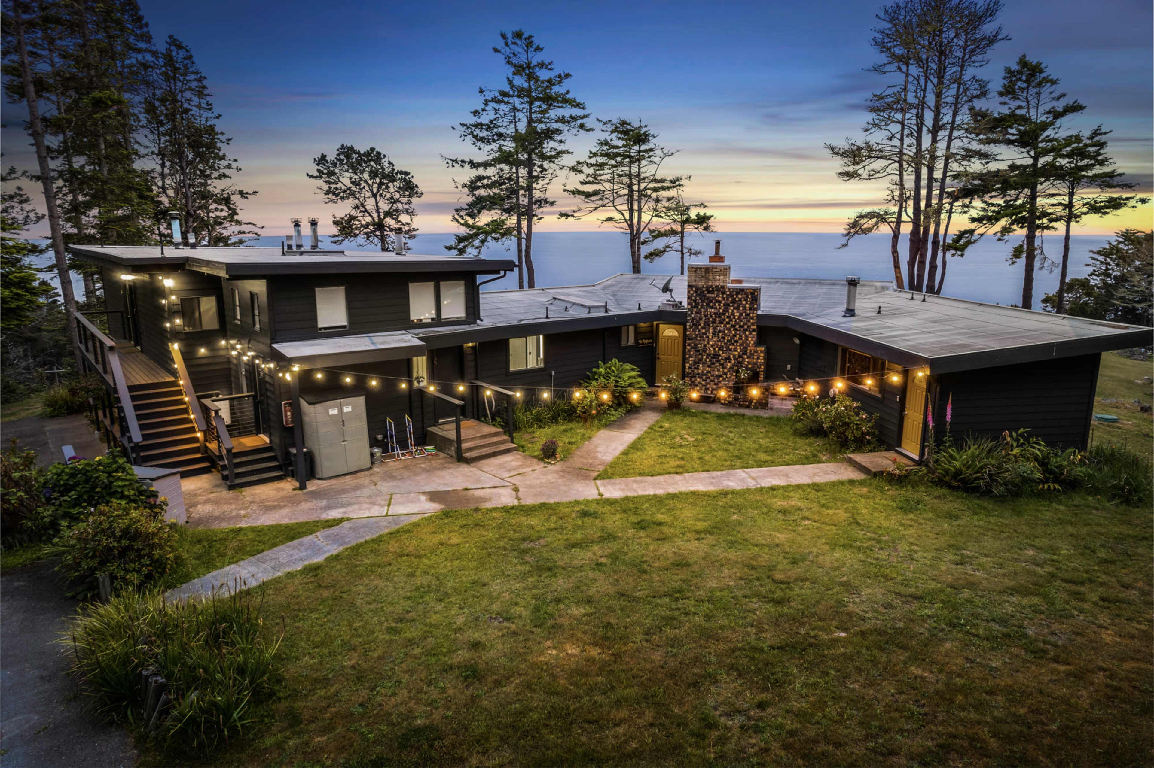 A modern black house with multiple levels and large windows is situated on a grassy lot overlooking the ocean at sunset, surrounded by tall trees.