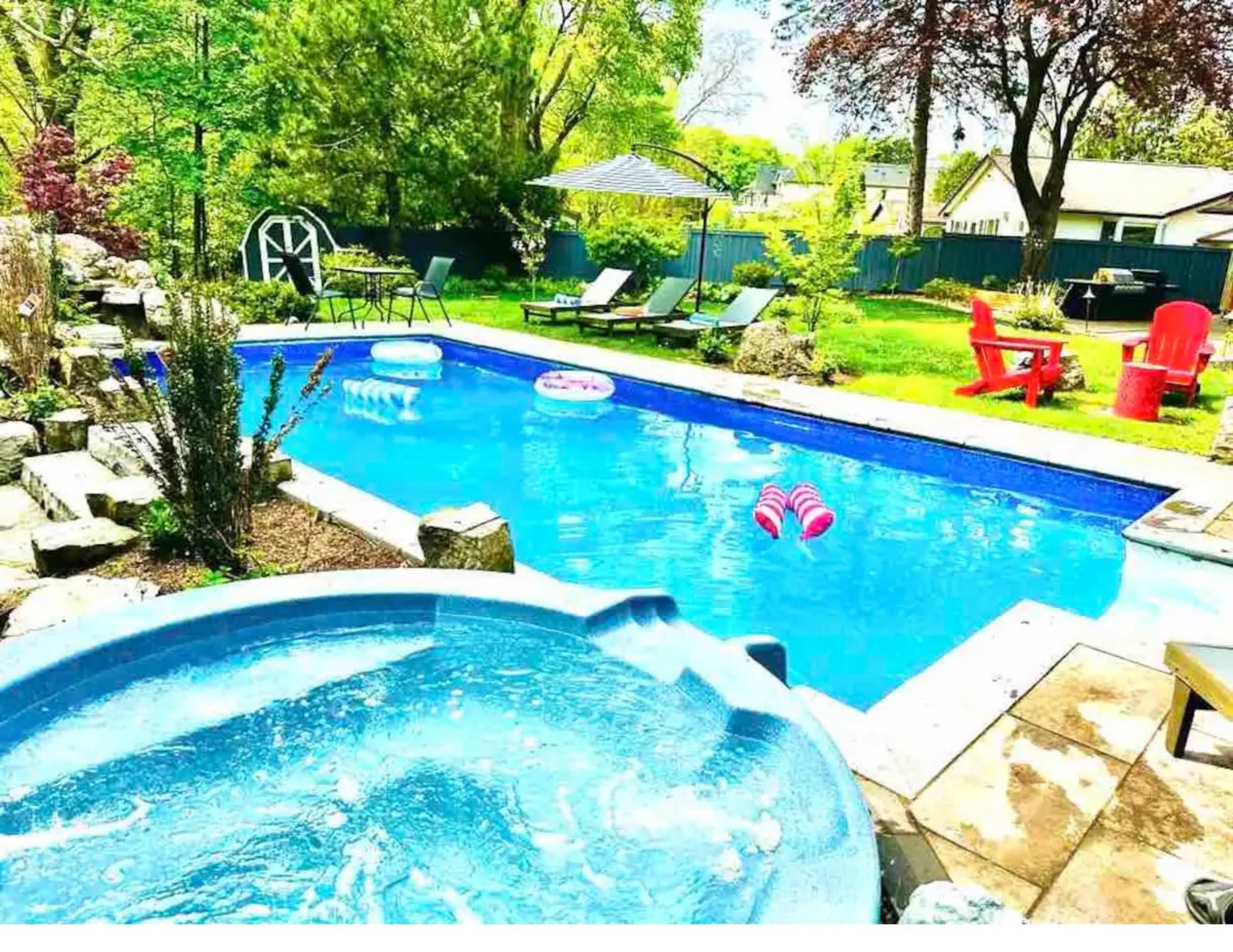A backyard pool area with a hot tub, lounge chairs, and colorful pool floats surrounded by greenery.