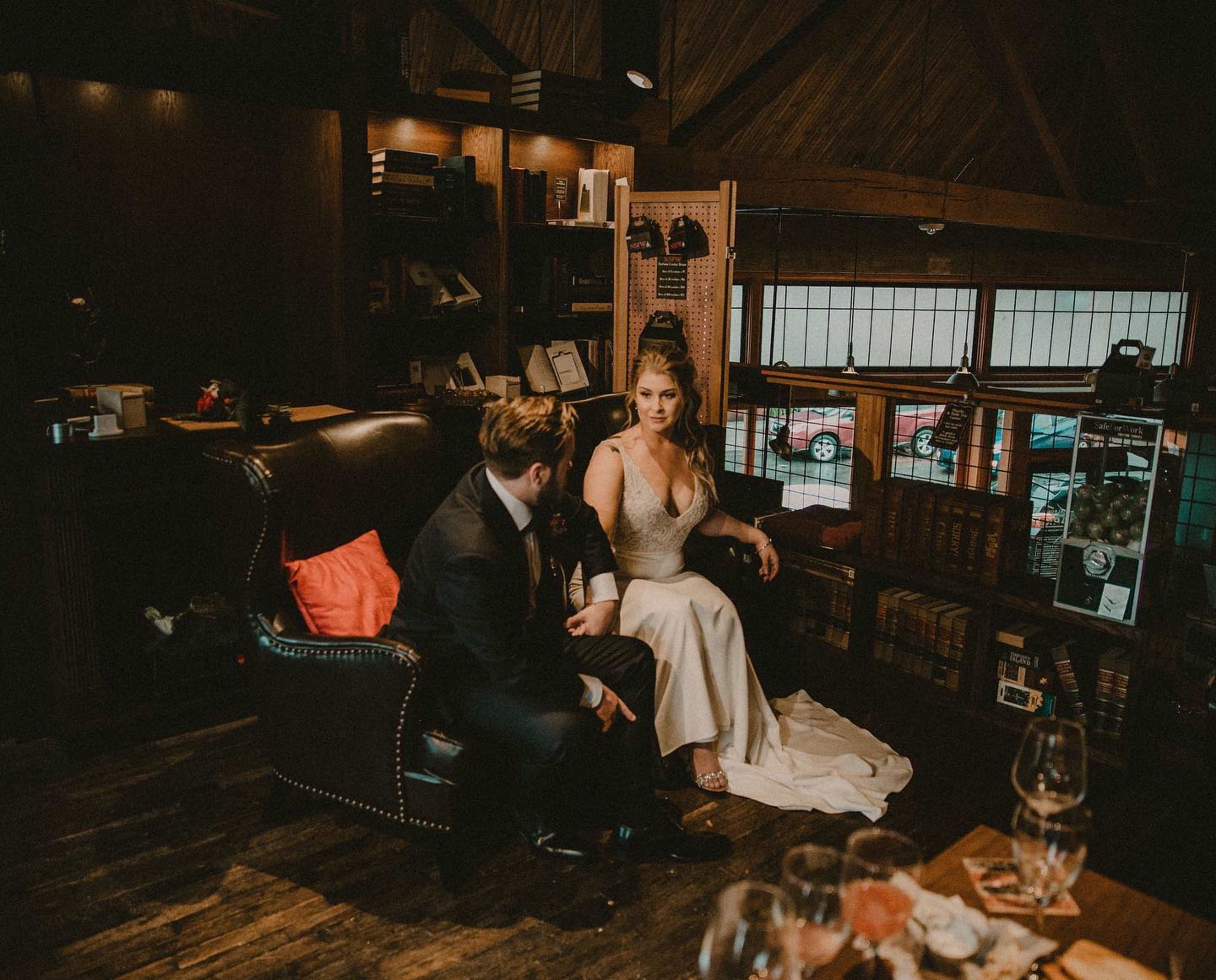 A couple sits together in an intimate setting, surrounded by bookshelves and warm lighting, with glasses on a table nearby.