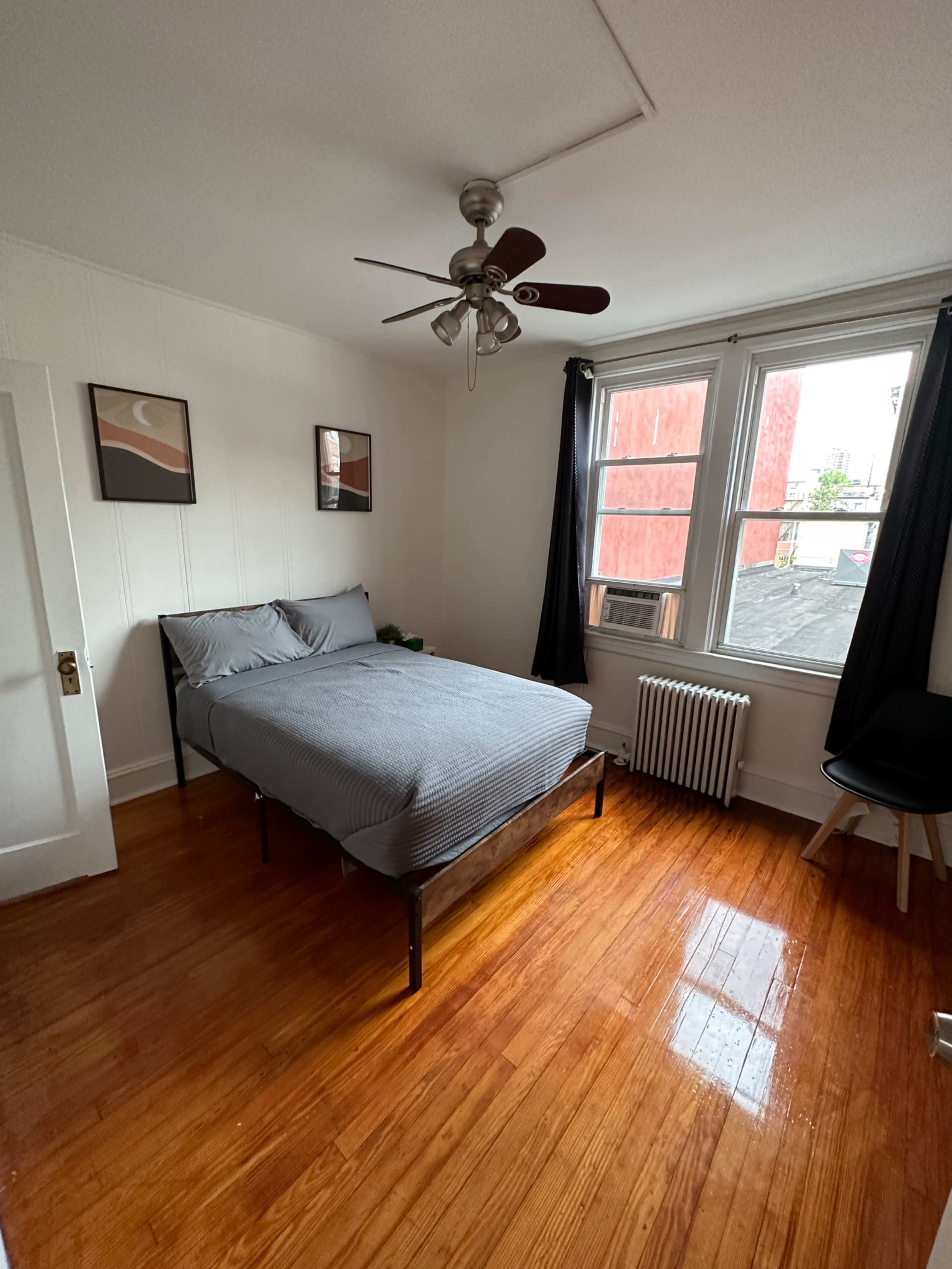 The room features a bed with gray bedding, a ceiling fan, and two framed pictures on the wall, with a window overlooking an exterior view.