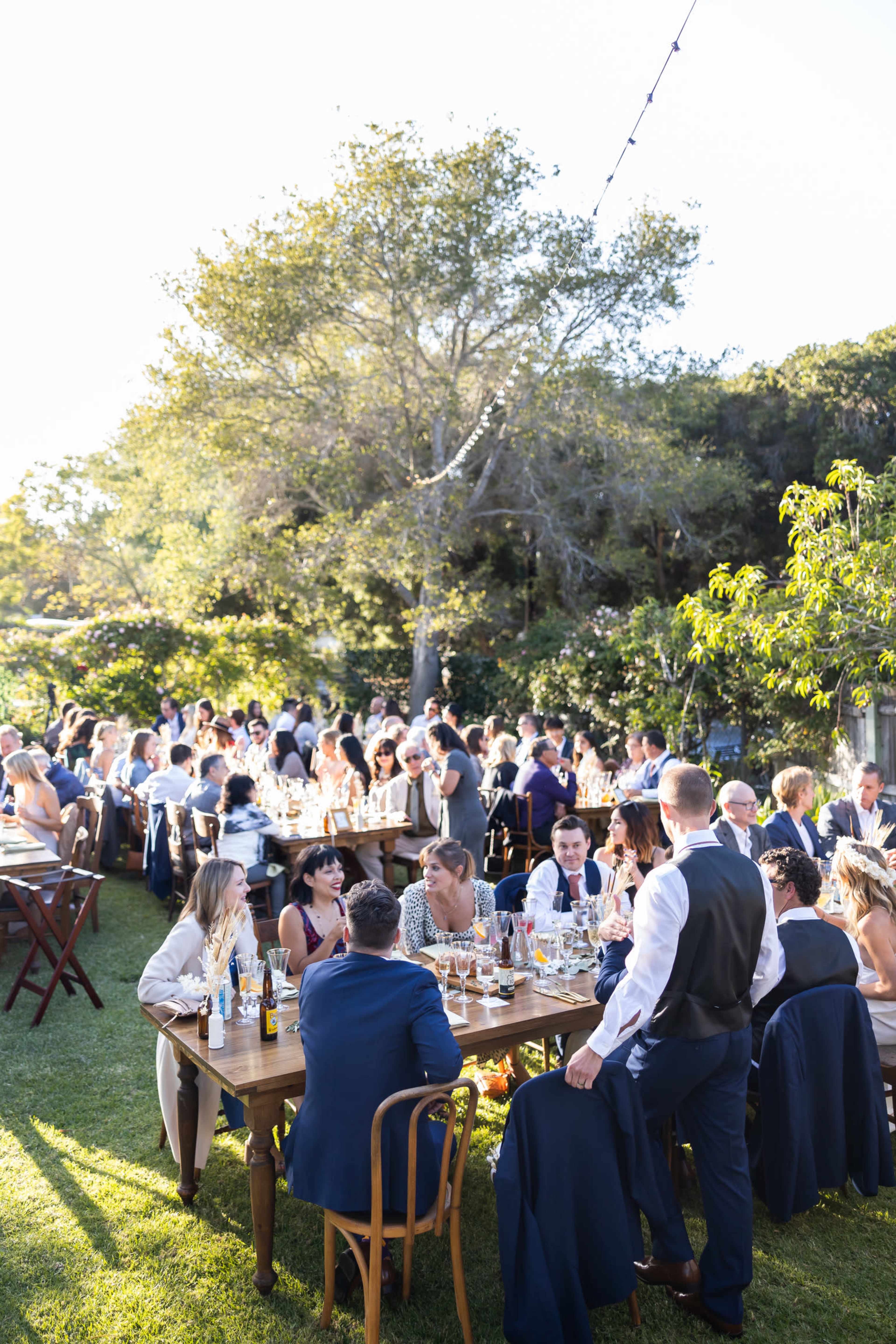 A large outdoor gathering features multiple tables filled with guests enjoying a meal under a sunny sky surrounded by greenery.